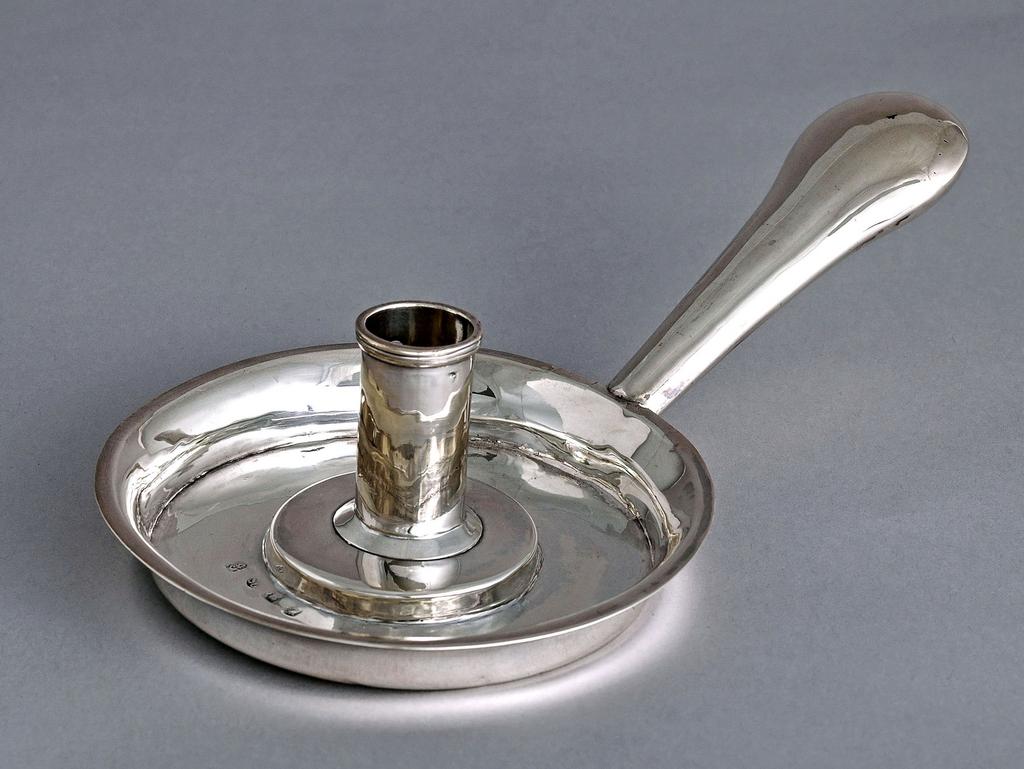 An image of Chamberstick. South, William, silversmith, London. Circular dish-shaped base with handle set at right angles to the base. The circular dish-shaped base with a raised centre supporting a cylindrical sconce. The side of the sconce is pierced with a long vertical bone-shaped hole, presumable to hold a mechanism for the raising of the candle. The long spatulate handle is hollow. Silver, height, to rim, 2 cm, width, handle to rim, 24 cm, diameter 13.5 cm, weight 250 g, 1705-1706. Queen Anne.