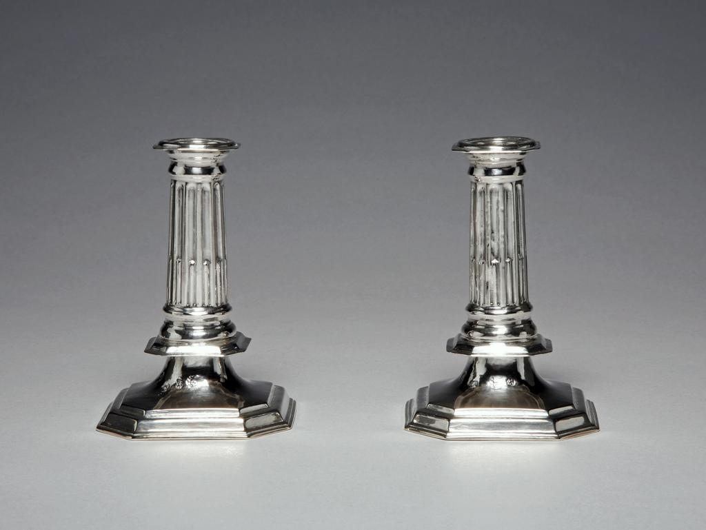 An image of A pair of miniature candlesticks. Ash, Thomas, silversmith, probably, London.M.39A-1961: A moulded octagonal rim above a sloping cylindrical column embossed with stop flutes on an octagonal knop above a trumpet-shaped foot on a stepped octagonal base. Silver, height, overall, 12 cm, weight 118 g, 1709-1710. Queen Anne.M.39B-1961: A moulded octagonal rim above a sloping cylindrical column embossed with stop flutes on an octagonal knop above a trumpet-shaped foot on a stepped octagonal base. Silver, height, overall, 12.2 cm, weight 124 g, 1709-1710. Queen Anne.