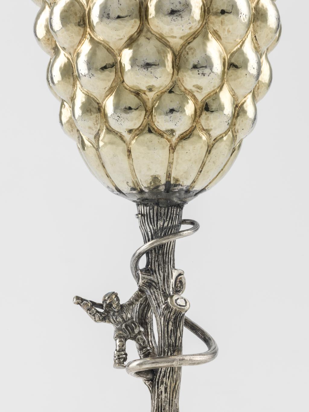 An image of Cup and cover/pineapple cup. Fischer, Franz (silversmith, Nüremberg, surname is also spelt Vischer). Lobed ovoid body and cover supported by a stem in the shape of a tree trunk with figure of a woodman. Silver-gilt; the spreading circular silver-gilt foot is embossed with a ring of lobes round a central dome, which supports the silver stem in the shape of a tree trunk encircled by a creeper, and the figure of an ax-wielding woodman. The ovoid silver-gilt body and interlocking cover are embossed with lobes. The silver finial is formed as a bunch of flowers. Silver and gold, raised and embossed body and foot, cast stem and woodcutter, height, overall, 33.3 cm, diameter, overall, 8 cm, weight, whole, 281 g, 1600-1653. Mannerist.