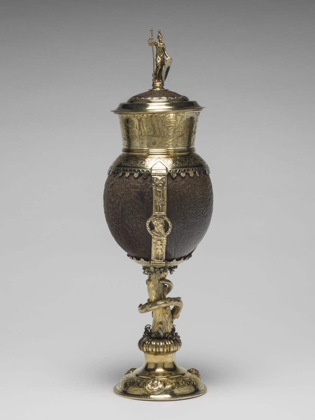 An image of Standing cup and cover. Unknown silversmith. The slightly domed circular base with three applied medallions of Classical heads between engravings of scrolling foliage and birds - including an owl. Between the base and the stem is a gadrooned knop topped with silver stylised leaves and scrolls. The stem is formed as a trunk of a tree with a branch spiralling round it. Above it is another ring of silver stylised leaves and scrolls and a calyx engraved with scrolls. Three straps contain the coconut. They are cast with a Classical figure standing in niche above and below a central medallion of the head of a bearded man surrounded by a laurel wreath. Around the top of the coconut is an border engraved with birds amid scrolling foliage, the edge pierced crockets. The deep rim mount is plain inside, whilst the exterior is engraved with a hunting scene showing two men on horseback with hounds chasing a stag and deer. The centre of the cover is of coconut shell with a silver-gilt border and central cast finial of a Classical female warrior. Coconut cup, silver-gilt mounts, gold surface of mounts, height, overall, 32.8 cm, width, overall, 10.5 cm, possibly 1560-1600. Probably German.