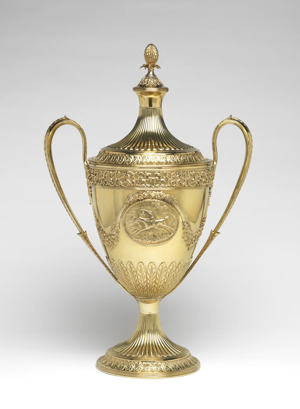 An image of Race Cup. Bateman, Peter. Bateman, Ann. Production Place:  London. The circular foot has a reeded edge above which is a band of leaf and bud ornament on a matted ground; the stem is embossed with bat's wing inverted fluting with an area of matting at the lower end of the flutes. The lower part of the tapering bowl has a calyx of lanceolate, wavy-edged leaves, above which, on each side of the body, is an oval medallion framed by husks. One medallion contains a plaque, cast and chased with two race horses and their jockeys racing in a landscape; the other is engraved with a coat of arms, sable a fess dancetty or in chief three fleur de lis argent impaling azure three uniorns' heads erased argent 2 and 1. Above each medallion there is an applied festoon of flowers and foliage from which depend two more festoons on either side, which loop up to meet the handles. The upper rim of the bowl has an applied frieze of strapwork and scrolling folliage within two moulded borders. The handles have lanceolate wavy-edged leaves at each upper end and a rib of husks on the exterior. The decoration of the cover follows that of the foot, but rises to a dome with reeded border topped by wavy edged leaves and an inverted pineapple finial. Decorated with cast and chased, applied, embossed and matted ornament and engraved with a coat of arms. Silver-gilt, height, overall, 59.9 cm, width, overall, 38.7 cm, 1799-1800. Neoclassical. Notes: The coat-of-arms on the dexter side are for Feeke, Feake or Feke of Wighton, Norfolk, or Hewster of Norfolk. The arms on the sinister side have not been identified, but could be for Shelley of Norfolk if the field was gules not azure.