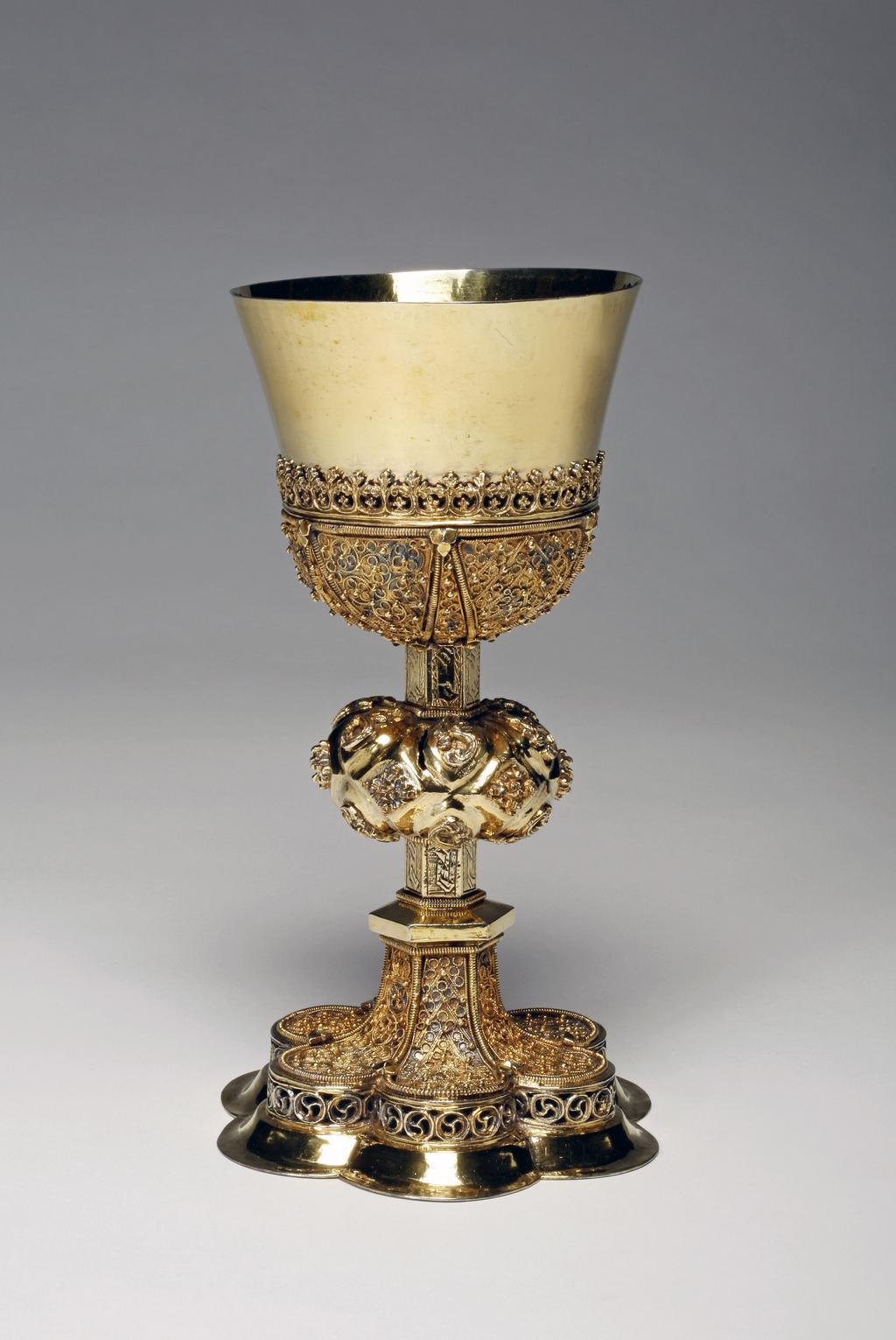 An image of Chalice. Unknown silversmith, Transylvania. Decorated with engraved, embossed, pierced, cast and filigree ornament. The base sits on a plain spreading hexafoil ring, above which is a verticle openwork border of cast scrolls. The trumpet-shaped foot is divided into six panels by ropework borders which contain applied filigree work. The hexagonal stem is engraved with a geometric pattern. At the centre is a large lobed knop with applied scrolls and flowerheads. The base of the plain U-shaped cup is held by triangular panels of filigree work, above which is a girdle of fleur-de-lys. Silver-gilt, raised, cast, applied, pierced and engraved, height, to rim, 20.7 cm, width, base, 11.7 cm, diameter, rim, 10.2 cm, weight 491 g, circa 1500-1550. Late Medieval.