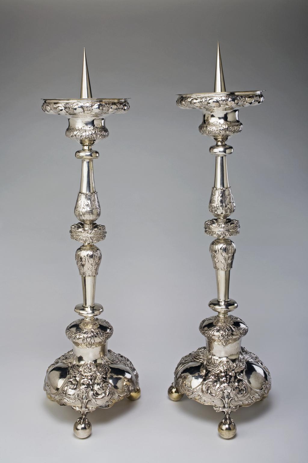 An image of One of a pair of altar candlesticks.MAR.M.76B-1912: Altar candlestick. Unknown silversmith, Netherlands, Dutch. The pricket candlestick embossed and chased with cherubs, gadroons and foliage. Only the pricket and drip pan are silver-gilt. Thse are supported on a bell-shaped sconce, which is embossed and chased with a border of gadroons and leaves. The stem comprises two balusters - one inverted - each with a plain knop at the outer end and an acanthus leaf embossed inner end, meeting in a flattened gadroon and leaf embossed and chased knop. The bell-shaped foot is embossed and chased with a border of swags, leaves and ribbons above three winged cherubs with vacant cartouches between. The three ball feet are each surmounted by a claw with leaf capital. Silver and silver-gilt, wooden base, height, overall, 74.3 cm, diameter, base, 20.2 cm, approximate, circa 1650-1700. Notes: The roundels of Christ are after plaquettes by an artist in the workshop of Matthias Wallbaum (1554-1632). MAR.M.76B-1912: Altar candlestick. Unknown silversmith, Netherlands, Dutch. The pricket candlestick embossed and chased with cherubs, gadroons and foliage. Only the pricket and drip pan are silver-gilt. Thse are supported on a bell-shaped sconce, which is embossed and chased with a border of gadroons and leaves. The stem comprises two balusters - one inverted - each with a plain knop at the outer end and an acanthus leaf embossed inner end, meeting in a flattened gadroon and leaf embossed and chased knop. The bell-shaped foot is embossed and chased with a border of swags, leaves and ribbons above three winged cherubs with vacant cartouches between. The three ball feet are each surmounted by a claw with leaf capital. Silver and silver-gilt, wooden base, height, overall, 75.2 cm, diameter, base, 20.2 cm, approximate, circa 1650-1700. Notes: The roundels of Christ are after plaquettes by an artist in the workshop of Matthias Wallbaum (1554-1632).