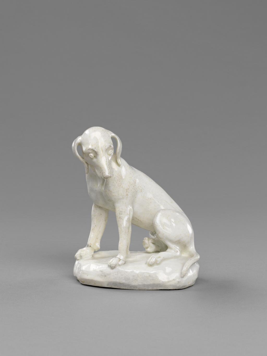 An image of Animal figure. Dismal Hound. Probably made by Charles Gouyn of St James's, London. The unglazed underside has a large oval opening, leaving a flange round the edge. The hound bitch sits on an oval, low mound base with its front paws apart and its head turnned to its left, looking downwards. It has floppy ears and a lugubrious expression. Soft-paste porcelain, slip-cast and lead-glazed. The glaze is off-white and very speckly. Height, whole, 12.5 cm, length, whole, 10 cm, circa 1749-1759. Rococo.