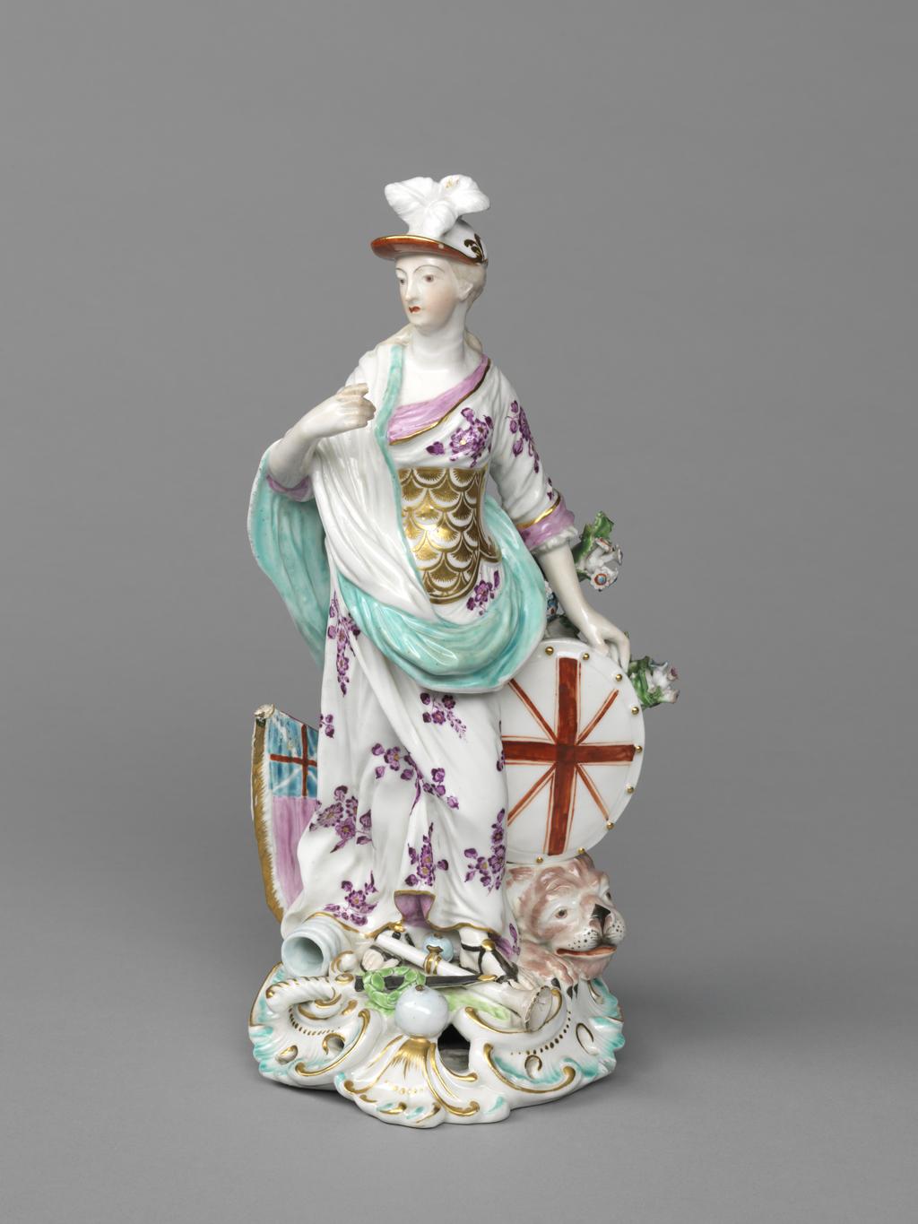 An image of Figure. Britannia. Derby Porcelain Factory, Derbyshire. William Duesbury & Co., proprietor. The base is approximately circular with a rococo scrolled and pierced front edge, and rises up at the back into a broad tree trunk with two branches bearing flowers and foliage on the figure’s left. Britannia stands with her left foot slightly advanced, her head turned a little to her right as she looks towards her raised right hand. Her left arm is a little behind her, the hand supporting an oval shield charged with red crosses of St George and St Andrew, which rests on the head of a lion. She has long, pale brown hair, tied back, and a cap with three white feathers at the front and a red underside to the peak. She wears a white dress with a puce floral pattern, pink cuffs, and pink drape round the neckline, a gilt scale cuirass, a white and turquoise shawl draped over her right shoulder and arm, and black sandals with a gold button on top. On the figure’s right a pink flag with a Union Jack in the top corner projects from the tree trunk. On the base there is a pale blue canon mouth, two pale blue canon balls of different sizes, a sword with a black scabbard with green wreath resting on it, and a white trumpet. The edge is picked out in turquoise and gold. Soft-paste porcelain, slip-cast, and painted overglaze in pale blue, turquoise, green, flesh pink, pink, puce, red, pale brown, and black enamels, and gilt. The unglazed underside has been ground flat, and has three patch marks, and a large circular central ventilation hole. Height, whole, 26 cm, width, whole, 12.8 cm, circa 1765. Rococo.