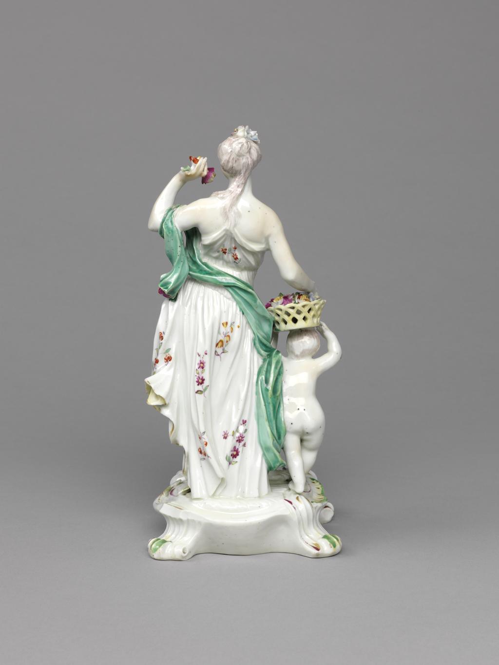 An image of Figure group. Spring (from a set of the Seasons). Derby Porcelain Factory, England. Eberlein, Johann Friedrich, modeller, after (German, 1696-1749). The circular base has four scroll feet. The edges are decorated with two rocaille 'frills' and a scroll, and the top is strewn with applied flowers. Spring stands on her left leg with her right leg relaxed behind her, leaning back slightly, and looking to her left . She holds up two flowers in her left hand, and has her right over an openwork basket filled with multi-coloured flowers which is held up by a nude infant standing beside her. She has long brown hair partly in a chignon and partly hanging down her back, and decorated at the front with three flowers. Her arms and breasts are bare, and below them she wears a white tunic with a floral pattern, a yellow lining, and gold edges, which is help up by a gold strap over her left shoulder. A turquoise-green scarf is draped over her left upper arm, across her back and round her right side.The child has brown hair and eyes, and a red lips. The edge of the base is picked out in green, yellow, dark pink and gold, and the flowers are variously dark pink, red and yellow, and blue and yellow. Soft-paste porcelain, slip-cast, with applied details, lead-glazed, and painted in a little pale blue, turquoise-green, green, yellow, flesh, dark pink, red, brown, and grey enamels, and lightly gilt. The glaze has many black speckles. The unglazed underside has a large circular ventilation hole, and four patchmarks. Height, whole, 23.5 cm, width, whole, 11.2 cm, circa 1765 -1770. Rococo. Production Note: The figure was based on a model which belonged to a set of Seasons modelled by J.F. Eberlein (1696-1749).