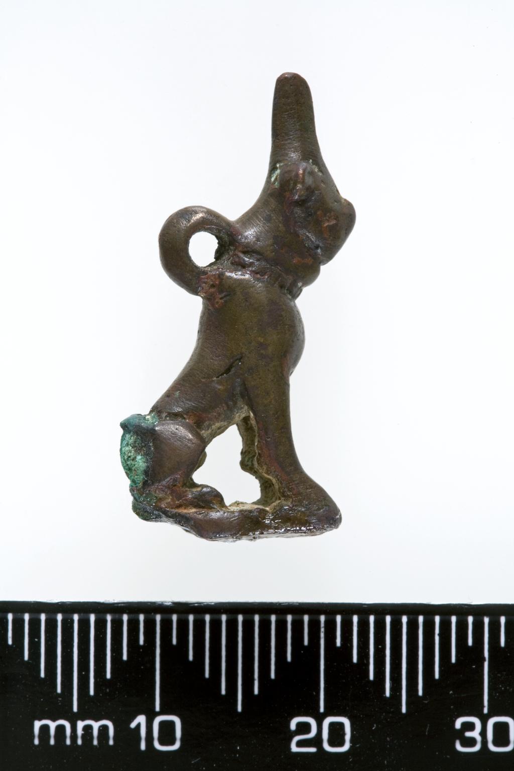 An image of Jewellery/Pendant. Dog pendant. Copper alloy (bronze), height 0.025 m, width 0.010 m, circa 600 B.C. Neo-Babylonian. Notes: Dating needs to be confirmed. Has been given date of 600 BC. However, may well be much earlier. e.g. Early Second Millennium.
