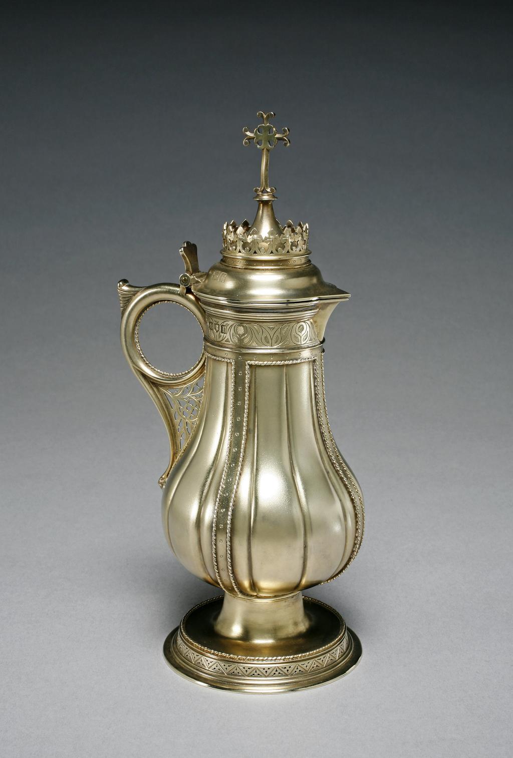 An image of Church Plate. Cruet. Keith & Co, London. The cruet has a baluster body and sparrow's beak spout, and stands on a stepped circular spreading foot. A border at the top of the body is engraved with a Gothic pattern of leaves, beneath which the body is fluted and devided into four panels by applied straps decorated with punched circles and a rope edge. The foot stands on a pierced gallery beneath an applied rope border. The slightly domed hinged cover is surmounted by a cross rising from a ring of trefoils. The handle is formed from a rope edged circle within a scroll, the area between being filled with a pierced grill of Gothic design. On the base is an inscription: To the Honour of God / and in memory of / Reginald Charles Coleridge / this Flagon was presented to / Hartford Parish Church / by his friends / many of whom were his comrades / in the Church Lad's Brigade / and the Boy Scouts / he went to his rest on the / 15 April 1912 / his body went down into the deep / with the wreck of the S.S. Titanic / and now sleeps in the hope / until the Sea shall / give up Her dead. Silver-gilt and matted, 1907-1908. Gothic Revival. On loan from Hartford Parish Church Council.