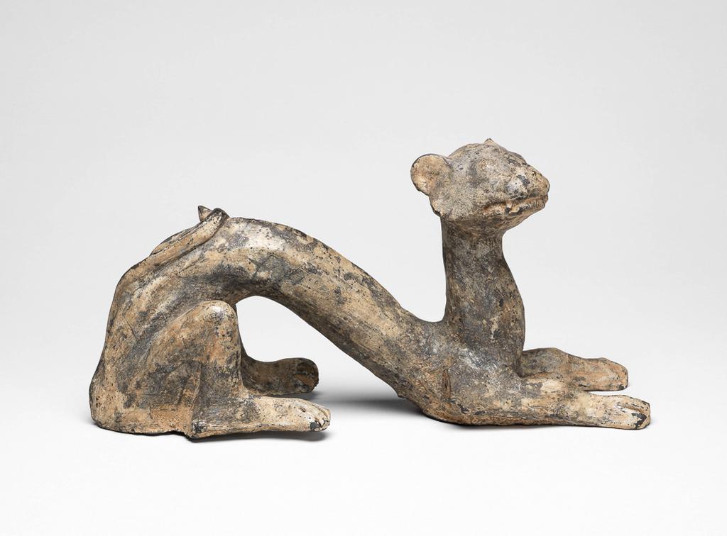An image of Figure. Crouching Feline Dragon. The dragon crouches with its back arched and its head up, its forelegs extended in front and its tailed curved up over its back. The wings, which would have projected from its back, are missing. Pottery, made from a pinkish buff fabric with uneven grey surface, height 13.9 cm, length 28.1 cm, 206 B.C. to AD 220. Han or Late Zhou. Chinese. Acquisition Credit: Lent to the museum by Helen H. L. Whittow (nee Malcolm).