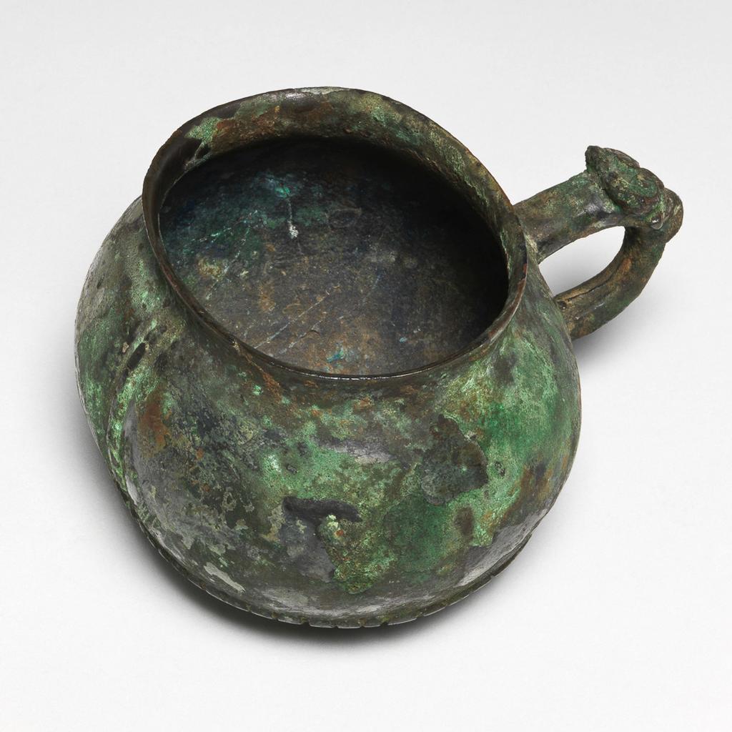An image of Drinking vessel. Bronze cup with a green patina on the exterior, and green and intense blue on the interior. Oval shaped with bulbous sides, slightly everted rim and loop handle with a bovine head on the upper part of its exterior. A short distance above the base, there is a horizontal seam simulating stiched leather, and on the side opposite to the handle, a vertical seam. Oval label printed in black with a tower and 'Malcolm Collection', inscribed in blue black ink 'A.7'. Circular inscribed in green '158'; rectangular printed in black 'Malcolm' and inscribed 'A.7'. Bronze, patina, height 7 cm, width 10 cm, 1050-771 B.C. Probably early Zhou (1050-771 BC). Chinese. Acquisition Credit: Lent to the museum by Helen H. L. Whittow (nee Malcolm).