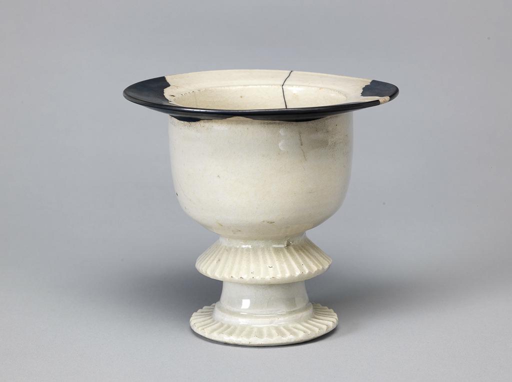 An image of Lamp or incense burner. The deep sided bowl has a wide, slightly sloping rim and is supported on a circular 'frilled' foot, and a cylindrical stem with a downward slanting 'frilled' flange at its junction with the bowl. Ding type. Two areas, one small one large, have been restored with what may be lacquer, and a crack has been filled with the same material. There are two short cracks in the rim, and six shallow chips on the underside of the foot. Porcelain or stoneware, with ivory white glaze and lacquer repairs, height 12.6 cm, diameter 13.5 cm, 1000-1127. Northern Song (960-1127). Acquisition Credit: Lent to the museum by Helen H. L. Whittow (nee Malcolm).