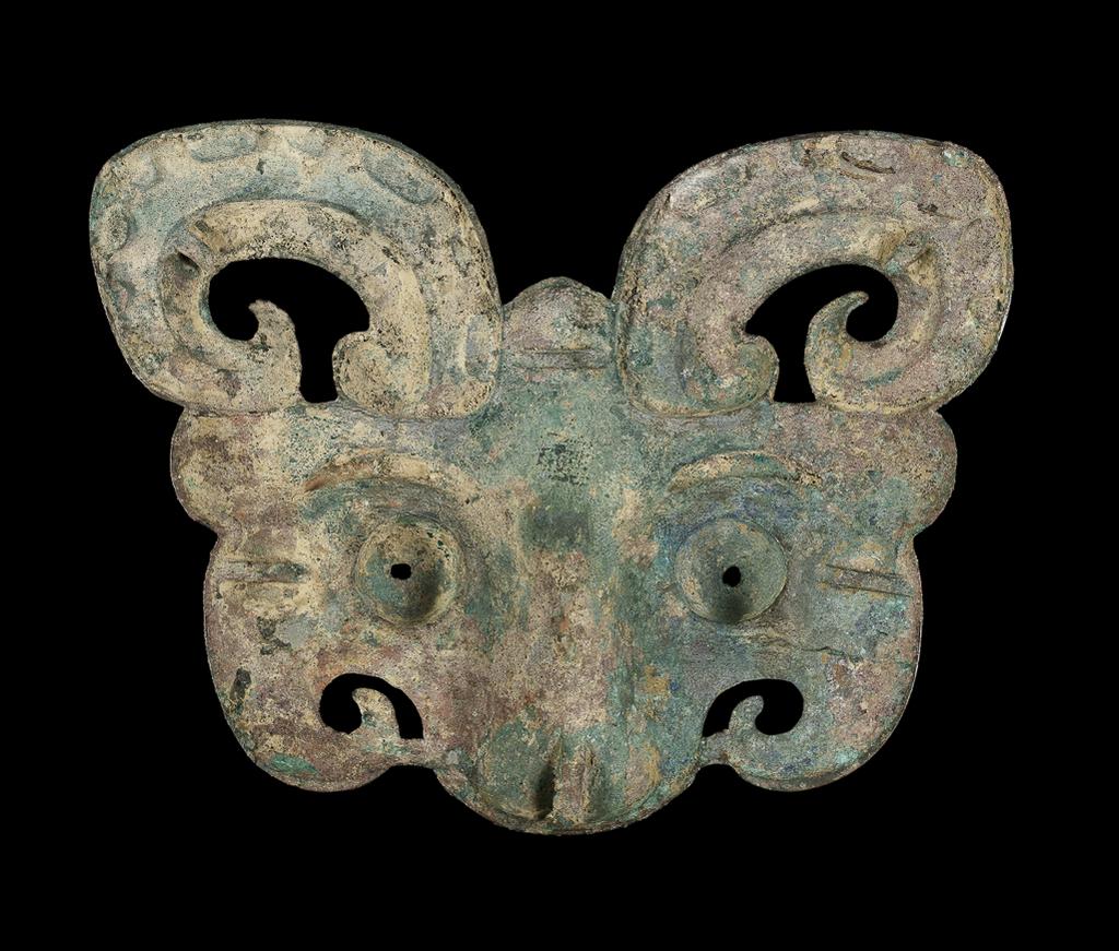 An image of Taotie mask. Feline face with curling horns, ridged eyebrows and pierced, bulging eyes, truncated at the tip of the nose. The back has six shallow loops for attachment. Label, with a tie on, inscribed in green ink 'Sir Neil Malcolm'. Bronze, with uneven green and reddish brown patination, pierced eyes, height 19.4 cm, width 25.5 cm. Zhou Dynasty (1050-771 BC). Chinese. Acquisition Credit: Lent to the museum by Helen H. L. Whittow (nee Malcolm).