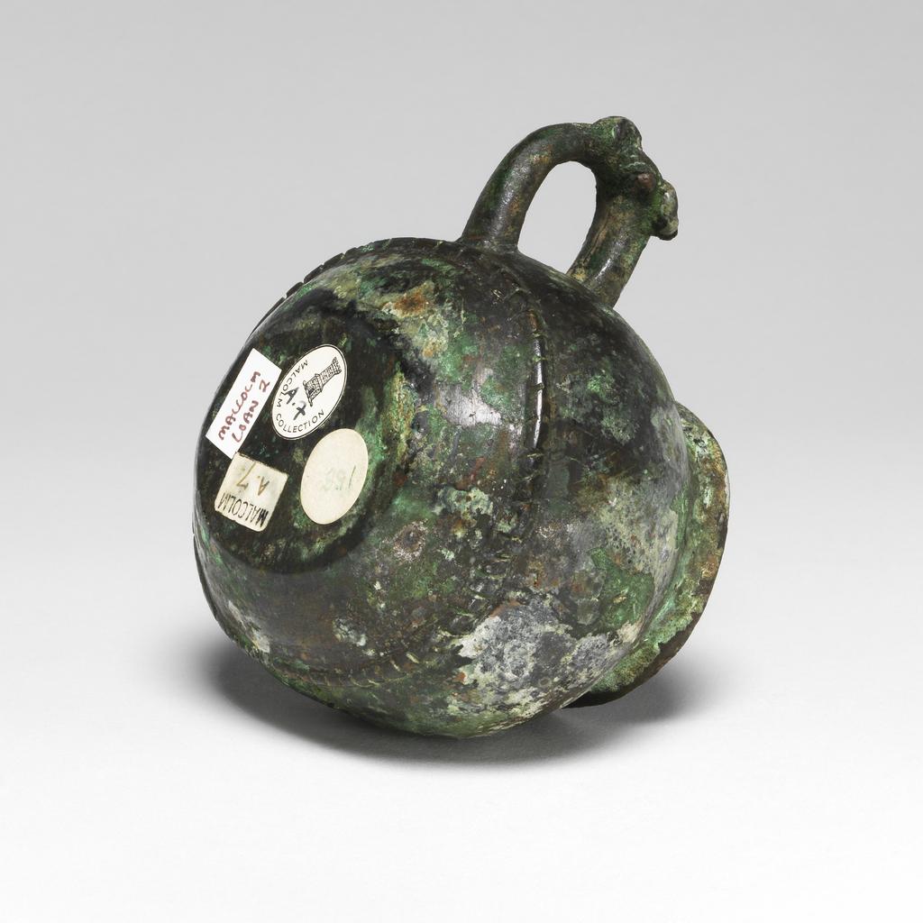 An image of Drinking vessel. Bronze cup with a green patina on the exterior, and green and intense blue on the interior. Oval shaped with bulbous sides, slightly everted rim and loop handle with a bovine head on the upper part of its exterior. A short distance above the base, there is a horizontal seam simulating stiched leather, and on the side opposite to the handle, a vertical seam. Oval label printed in black with a tower and 'Malcolm Collection', inscribed in blue black ink 'A.7'. Circular inscribed in green '158'; rectangular printed in black 'Malcolm' and inscribed 'A.7'. Bronze, patina, height 7 cm, width 10 cm, 1050-771 B.C. Probably early Zhou (1050-771 BC). Chinese. Acquisition Credit: Lent to the museum by Helen H. L. Whittow (nee Malcolm).