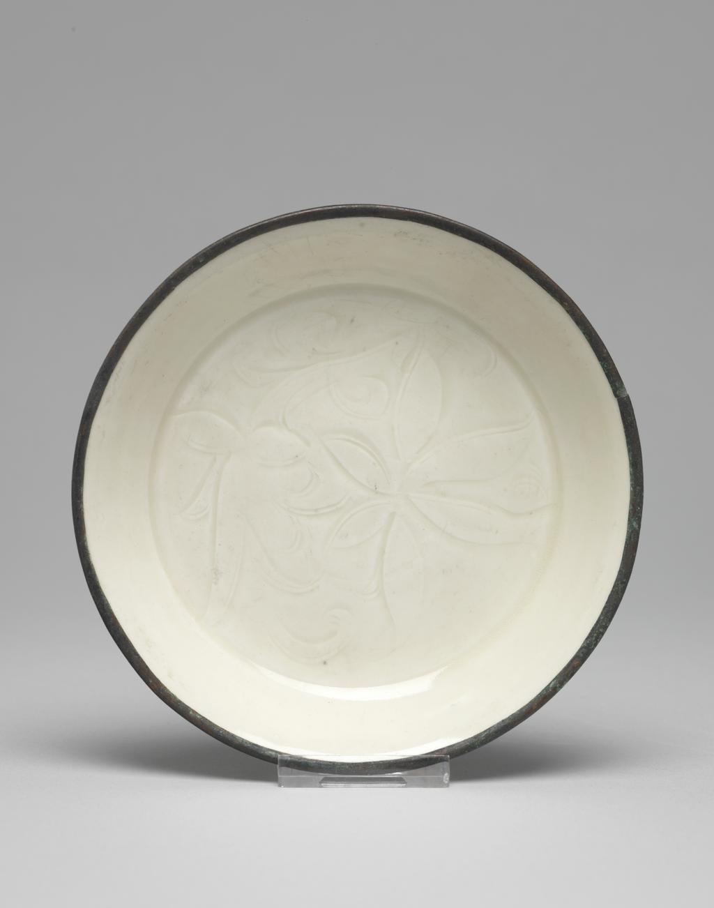 An image of Saucer. Ding Yao ware. The saucer is circular with shallow sloping sides. The flat centre is incised with a lotus flower and foliage. With an oval label printed in black, with a tower and 'Malcolm Collection', and inscribed in blue black ink with 'D./49'. White stoneware, with an incised decoration under the ivory white glaze, and a rim mounted in what may be copper. Height 2.5 cm, diameter 13 cm, 960- 1127. Northern Song (960-1127). Chinese. Acquisition Credit: Lent to the museum by Helen H. L. Whittow (nee Malcolm).