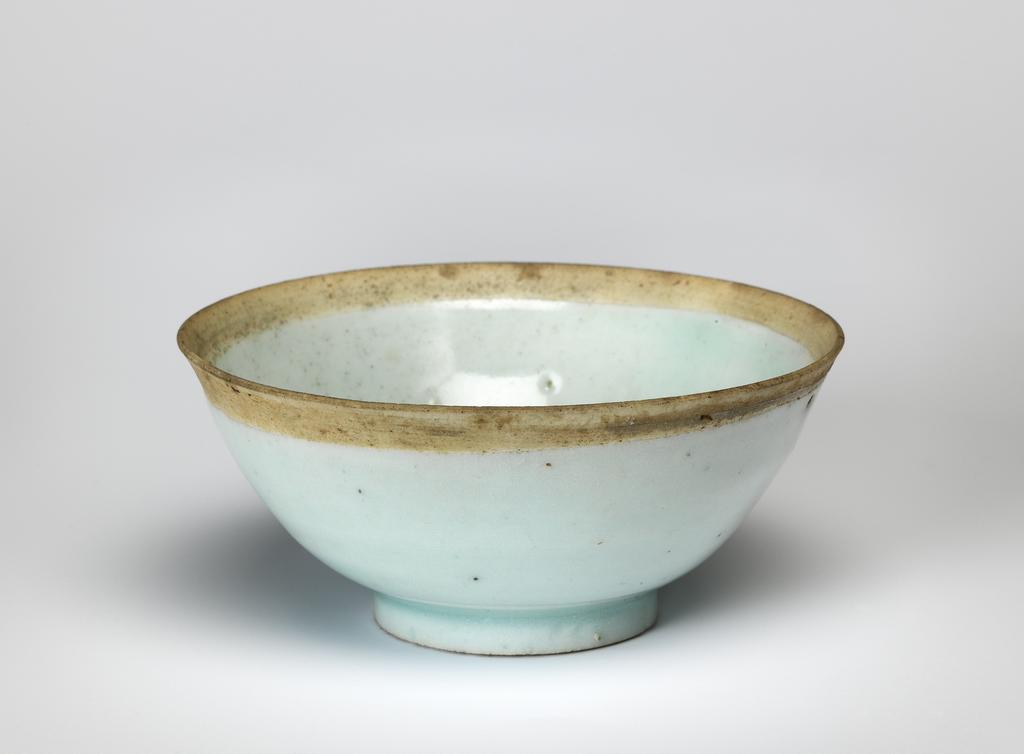 An image of Bowl. Qingbai ware. Circular, with deep, gently curving sides, standing on a footring. The glaze inside the bowl is speckled with black, particularly in the centre. Oval label printed in black with a tower and 'Malcolm Collection' and inscribed in blue black ink 'D./36'. Porcelain covered with very pale greenish blue glaze, except for approximately 7 cm on both sides of the rim. Height 4.7 cm, diameter 10.5 cm, 1100-1299. Song Dynasty (960-1279). Chinese. Acquisition Credit: Lent to the museum by Helen H. L. Whittow (nee Malcolm).