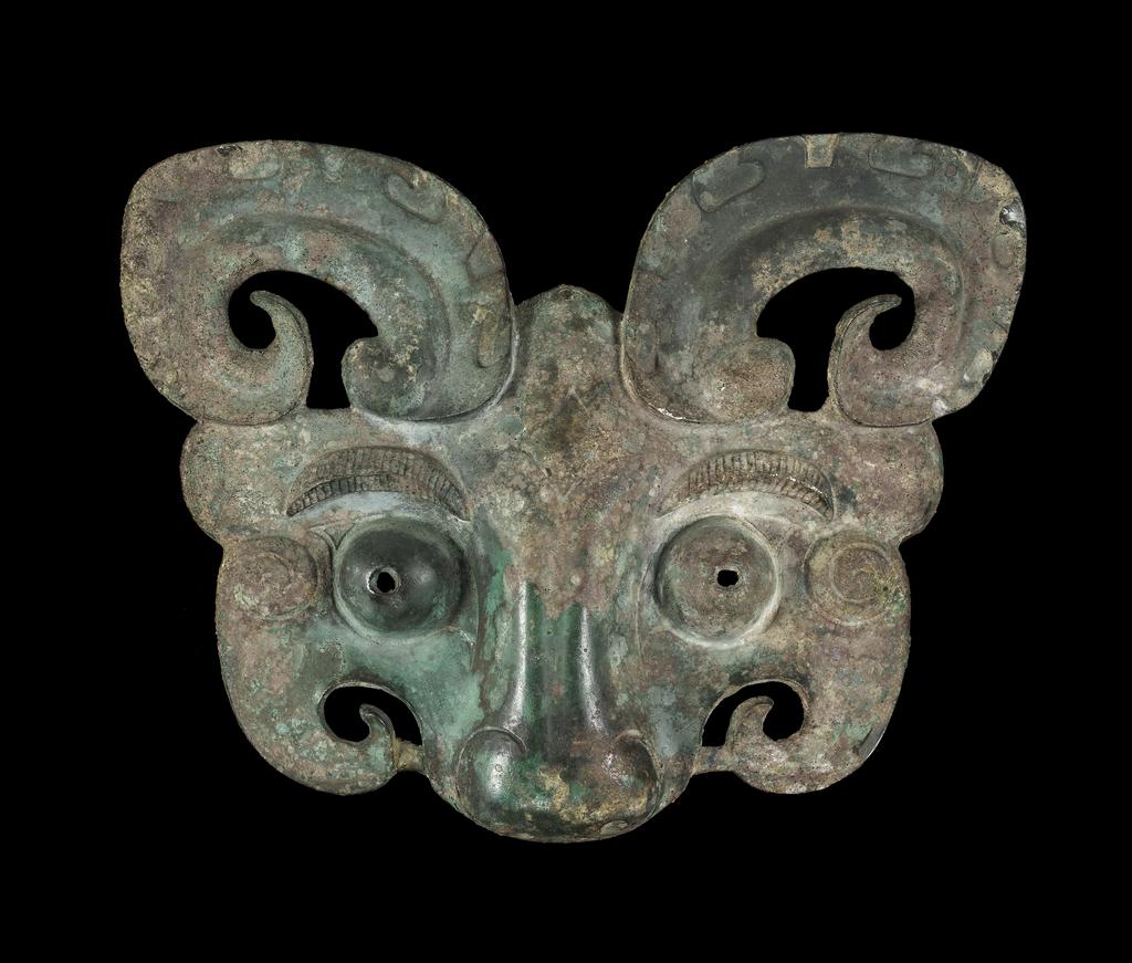 An image of Taotie mask. Feline face with curling horns, ridged eyebrows and pierced, bulging eyes, truncated at the tip of the nose. The back has six shallow loops for attachment. Label, with a tie on, inscribed in green ink 'Sir Neil Malcolm'. Bronze, with uneven green and reddish brown patination, pierced eyes, height 19.4 cm, width 25.5 cm. Zhou Dynasty (1050-771 BC). Chinese. Acquisition Credit: Lent to the museum by Helen H. L. Whittow (nee Malcolm).