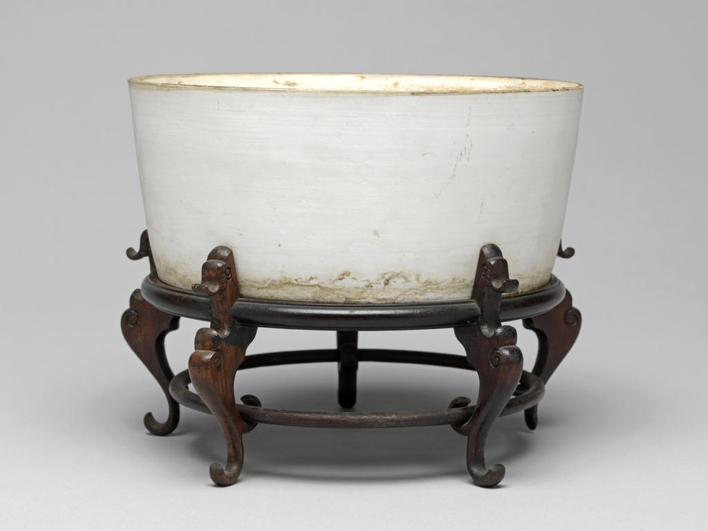 An image of Flower pot and stand. Circular with deep almost straight sides and slightly convex base, pierced by five drainage holes. Ding yao type. With an oval label printed in black with a tower and 'Malcolm Collection', and inscribed in blue ink D. 134. Circular wooden stand with five legs.There are brown stains around the interior, especially below the rim, and round the exterior near the base. A few stains and marks on the base. One leg of the stand is damaged on the inside. White unglazed porcelain, pierced base, height 7.4 cm, diameter 16.3 cm, after 960. Song Dynasty or later. Chinese. Acquisition Credit: Lent to the museum by Helen H. L. Whittow (nee Malcolm).