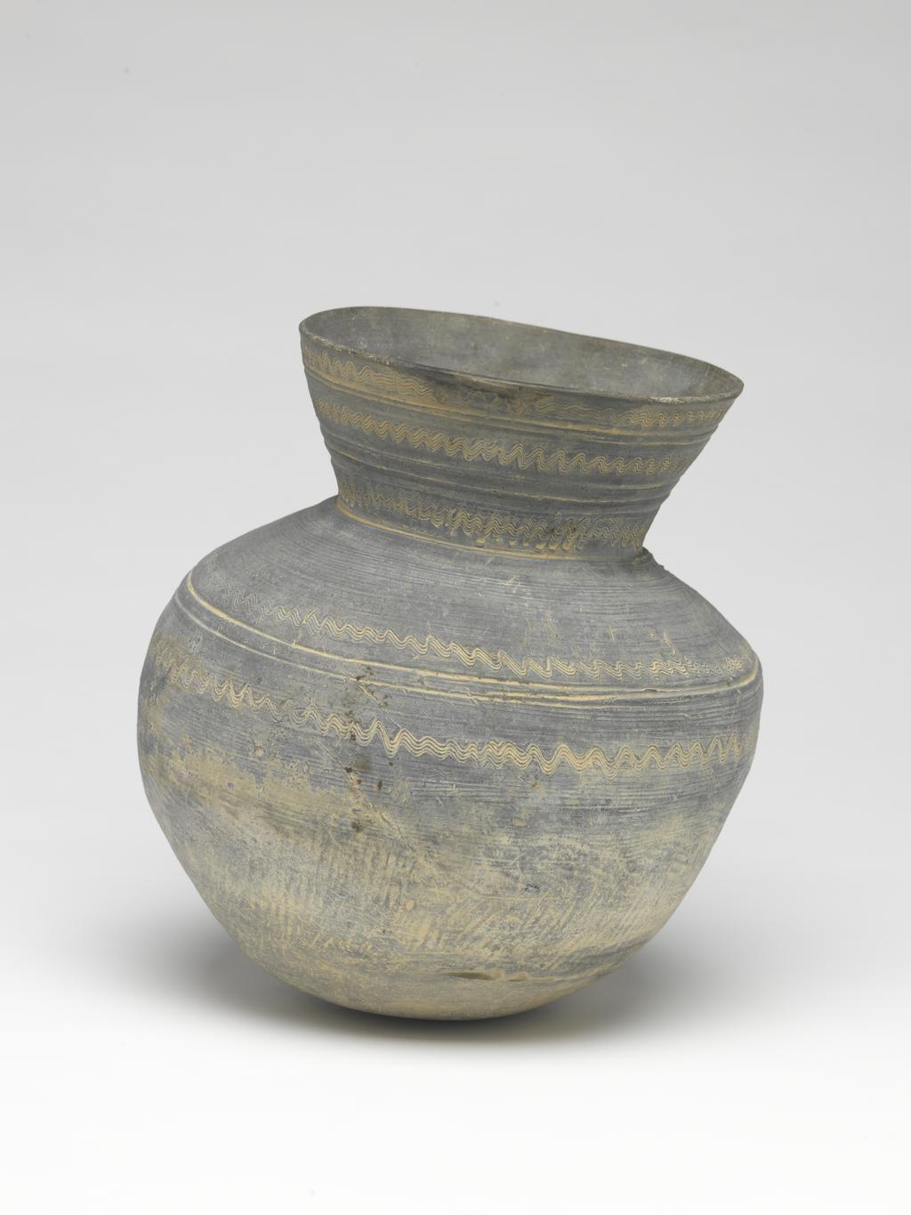 An image of Vase. Production Place: South West Korea. The globular body has a rounded base and straight sided flaring neck. The lower part of the body is decorated with faint impressed diagonal lines inclined in different directions. The upper part of the body and neck are smooth. The upper part of the body is decorated with a pair of incised horizontal lines between groups of close set wavy lines. On the neck there is a horizontal line, a group of close set wavy lines, three horizontal lines, and two horizontal lines between groups of close set wavy lines. There is a repair in the rim, and two dark shiny splodges over it. Hand built dark grey stoneware, incised decoration, height, 22, cm, 18 B.C. to AD 663. Paekche Kingdom. Korean. Acquisition Credit: Lent to the museum by Helen H. L. Whittow (nee Malcolm).