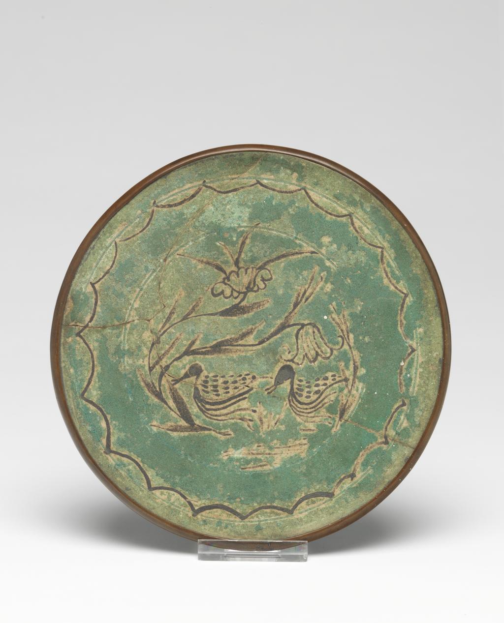 An image of Plaque. Cizhou type. In the middle there are two ducks on a pond, with a flowering plant to the left, surrounded by a circular scalloped border. The edge is mounted in copper. Oval label printed in black with a tower and 'Malcom Collection' and inscribed in blue-black ink 'D./63'. Circular inscribed in blue black ink '146'. Stoneware, decorated with painting in brown and turquoise green glaze. Reconstructed from three pieces. The glaze on both sides is degraded. Diameter 14.2 cm. Yuan Dynasty (1279-1368). Chinese. Acquisition Credit: Lent to the museum by Helen H. L. Whittow (nee Malcolm).