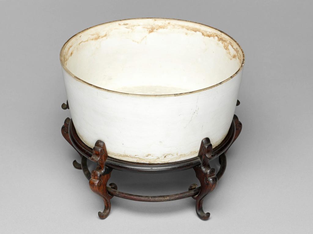 An image of Flower pot and stand. Circular with deep almost straight sides and slightly convex base, pierced by five drainage holes. Ding yao type. With an oval label printed in black with a tower and 'Malcolm Collection', and inscribed in blue ink D. 134. Circular wooden stand with five legs.There are brown stains around the interior, especially below the rim, and round the exterior near the base. A few stains and marks on the base. One leg of the stand is damaged on the inside. White unglazed porcelain, pierced base, height 7.4 cm, diameter 16.3 cm, after 960. Song Dynasty or later. Chinese. Acquisition Credit: Lent to the museum by Helen H. L. Whittow (nee Malcolm).