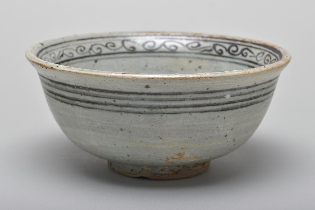 An image of Studio Ceramics. Stoneware bowl with underglaze iron decoration featuring an unusual phoenix motif in a central medallion with foot. Production Place: Thailand. Light grey-green glaze with iron spots, pin-holing, and fine crazing. Five spur marks in the well. Signs of cylindrical kiln support on the base, indicating that this bowl was at the bottom of a stack. 1300-1399. Acquisition: Dr John Shakeshaft Bequest.