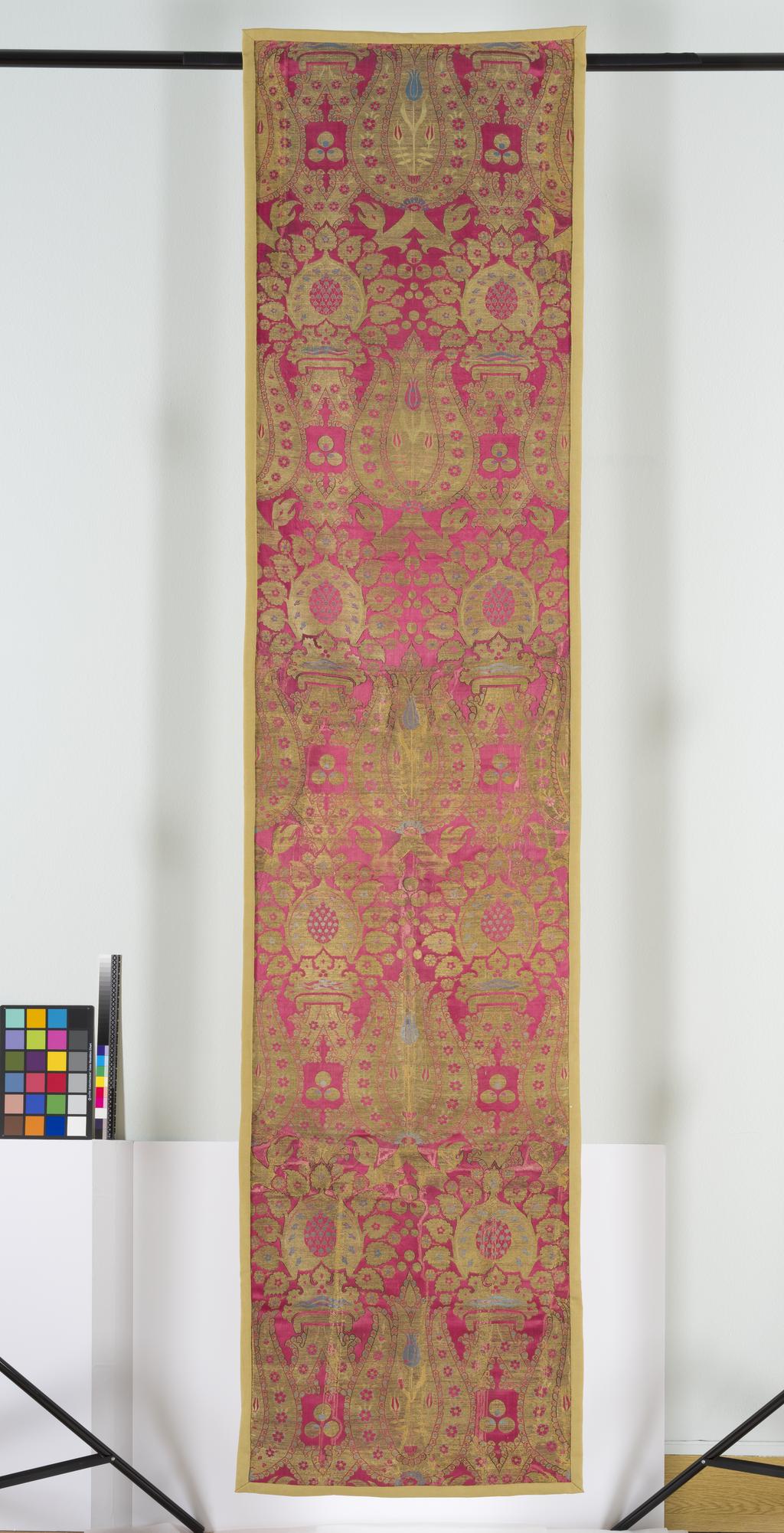 An image of Textiles. Panel. Unknown maker, Turkey. Lampas, red warp-faced satin ground with pattern formed by silk wefts of light blue and yellow with fragments of the black originally used for outlining the large areas of gilt metal thread, now largely worn down to its silk core. Ogival lattice tied by Italian crown motif with an alternating repeat pattern of tulip and artichoke. Length, whole, 290 cm, width, whole, 67.2 cm, circa 1550 to circa 1599.