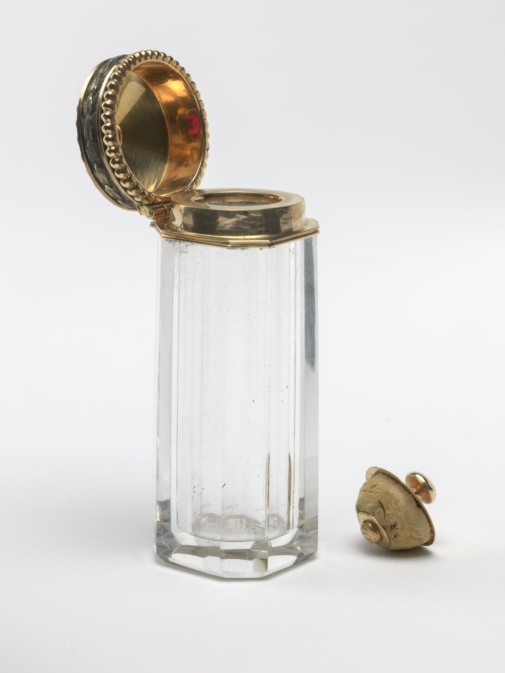 An image of Objets de vertu. Scent bottle/scent flask. Russian. The rock crystal body is of octagonal section with straight sides externally, and cylindrical internally; it has a gold rim mount and hinged lid. The lid has a border of beads round the rim, above which is a chased border of overlapping leaves enamelled in dark translucent green. The top of the lid is slightly domed and is embossed with flutes centering on a cabuchon garnet. Inside is a flat gold stopper (A) with compressed ball-shaped knop and cork base. With fitted case (B). Rock crystal perfume container, gold, enamel and a garnet, height, whole, 2.1, cm, length, whole, 6.0, cm, width, whole, 2.1, cm, circa 1900 to circa 1910.
