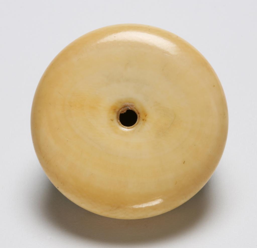 An image of Netsuke: kagamibuta. Unknown maker, Japan. Circular bowl, plain with indented ridge around the top edge and traces of green staining to inner rim, central himotoshi. Shibuichi disc, cast and decorated in relief with a fly resting on top of a man's shaved head above the wrinkled brow, the face cut off at the nose. The man's hand is raised as if to swat the fly. Finely stranded hair and eyebrows in shakudo and copper, the man's eyeballs in shakudo and copper, and the fly's eye is applied with copper. Ivory bowl with cast, metal disc decorated in relief and eyelet. Height, whole, 1.8 cm, width, whole, 4.2 cm, circa 1800-1868.
