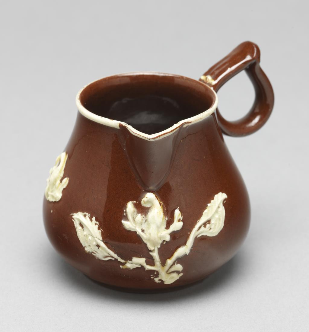 An image of Cream jug/milk jug. Unidentified Staffordshire Pottery, England. The bulbous body rises into a short cylindrical neck with a sparrow beak lip, and at right angles to it, a ring handle pinched together at the top. Below the lip is a sprig of bird perching on an upright branch with a large leaf on either side. Further to the left is a smaller bird perching on a branch with leaves, and to the left of that, a fleur-de-lys. A white slip band runs round the rim. Red earthenware, thrown, with applied spout and handle, decorated with mould-applied white sprigs (reliefs) and white slip under lead-glaze. Height, whole, 7 cm, width, whole, 8.4 cm, circa 1740-1750. Rococo. Sir Ivor and Lady Batchelor Bequest.