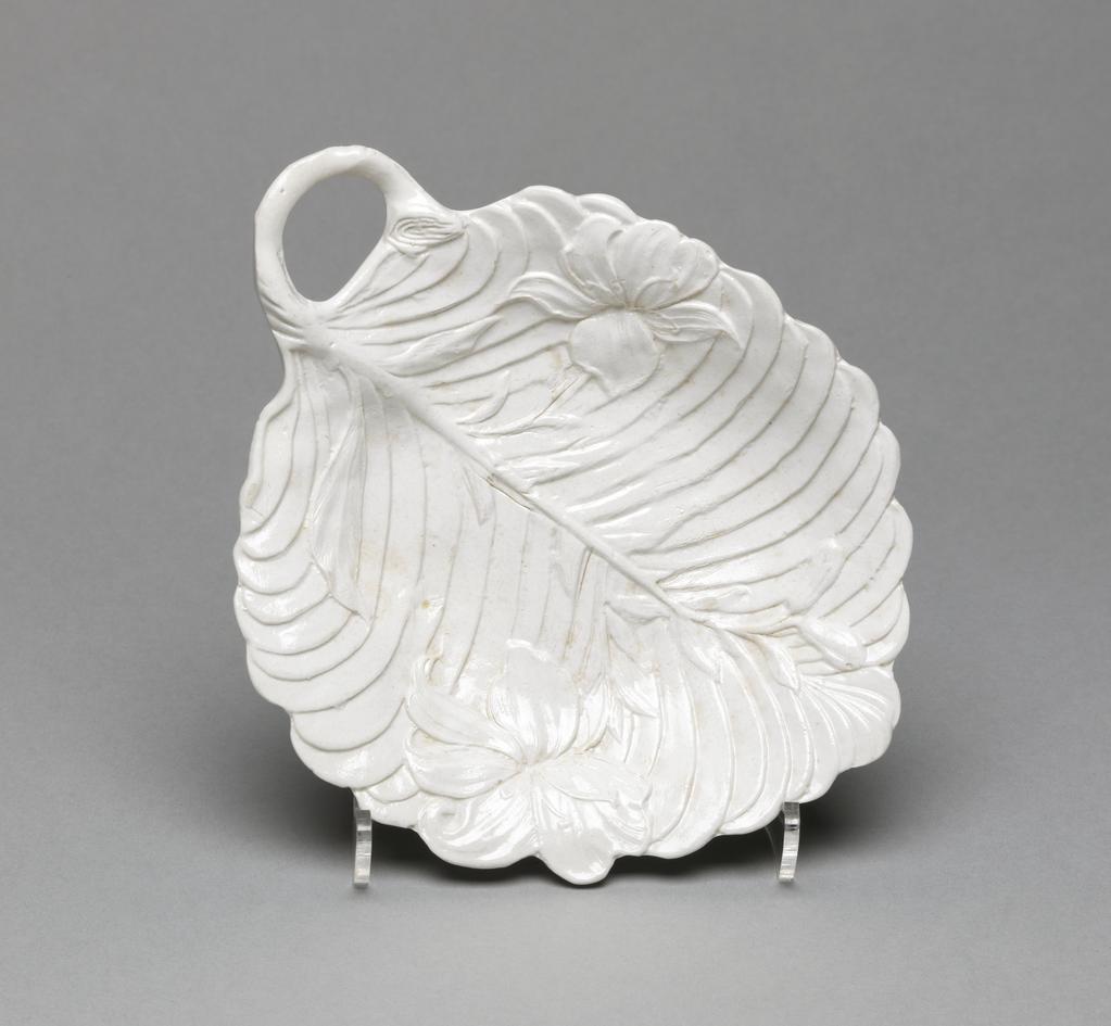 An image of Leaf Dish. Pickle Dish. Unidentified Staffordshire factory, England. In the shape of a concave leaf with a scalloped edge and bent stalk handle, standing on three fluted flower-shaped feet. The interior is decorated with a lily plant with one flower on each side and and three buds at the top, superimposed over the slanting veins of the leaf. Off-white stoneware, press-moulded with integral decoration, and salt-glazed. 1750-1770. Rococo. Sir Ivor and Lady Batchelor Bequest.