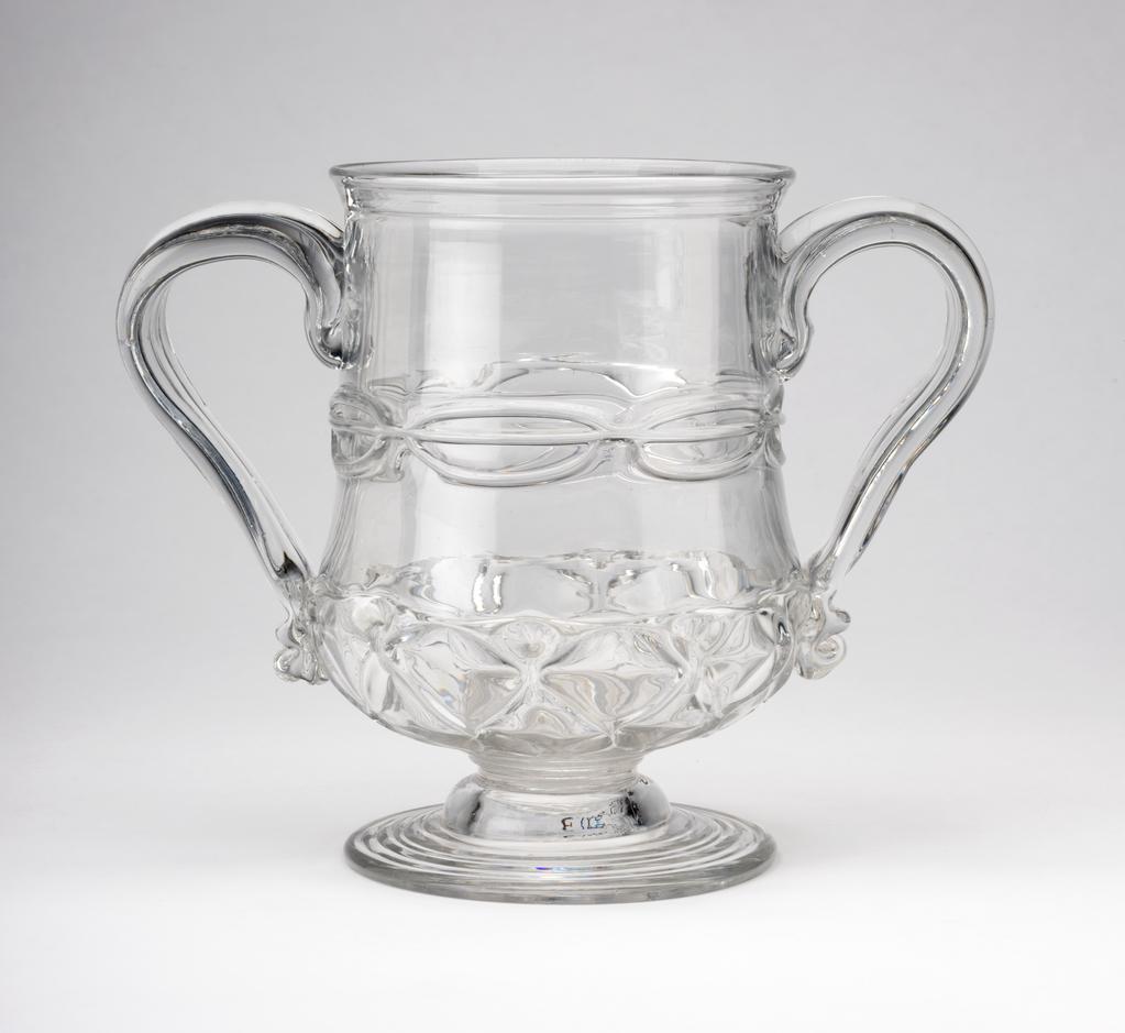 An image of Two-handled cup. Unidentified English glasshouse. The cup stands on a circular foot with a rough pontil mark underneath. The body is thistle-shaped with two applied loop handles.  The foot has indented rings. The lower part of the body is moulded with ribs, pincered into a repeating diamond pattern (nipt diamond-waies). The waist has three horizontally trailed threads pincered twice on each side and once under each handle to form a chain. The handles have a vertical ridge and a pincered kick at the bottom. Colourless lead-glass blown, pattern-moulded, pincered, and trailed. height, whole, 14.8 cm, width, whole, 20 cm, 1690-1710. Baroque. Sir Ivor and Lady Batchelor Bequest through The Art Fund.