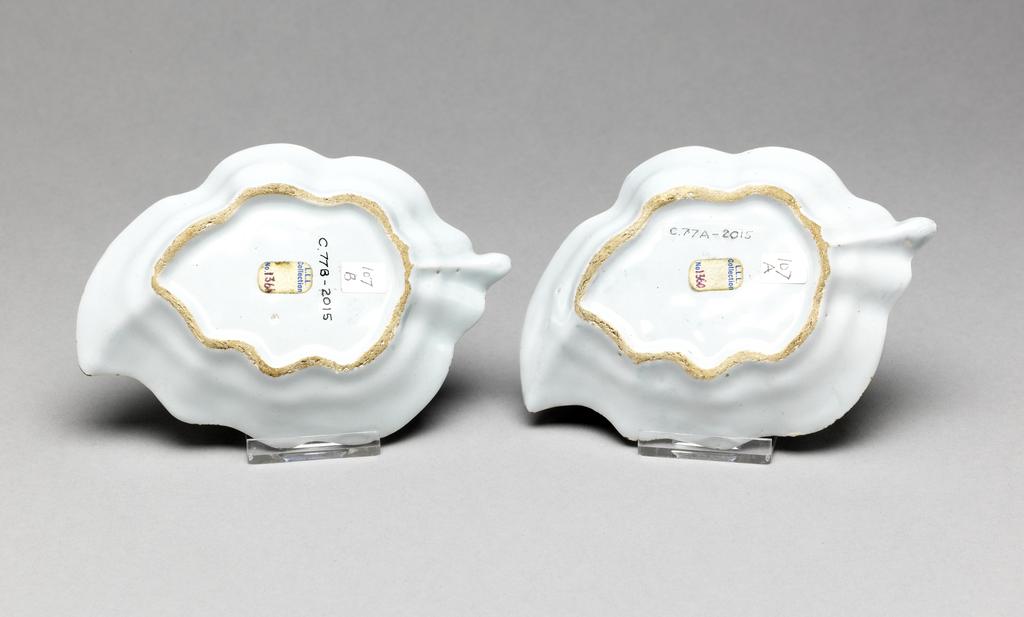 An image of English Delftware. Leaf dish. Pickle dish. Unidentified Liverpool pottery, England. Both dishes are moulded in the shape of a leaf with wavy edges and a short stalk, standing on a shaped footring. The interiors are painted with a lakeside scene in which there is a man in a boat in the foreground, an area with tufts of grass or reeds, three houses flanked by bushes and a single house with a fence to the right of it. Behind each house there is a slender tree resembling a telegraph pole, and overhead are three groups of schematic birds. The edge has a border of slanting lines broken by six groups of four spirals. Buff earthenware, press-moulded, tin-glazed pale bluish-white and painted in blue. Height, A, 2.2 cm, length, A, 13.2 cm, width, A, 9.2 cm; height, B, 2.2 cm, length, B, 13.1 cm, width, B, 9.3 cm, circa 1760. Rococo. Production Note: Small dishes of this shape were probably used for pickles presented on the table between the larger dishes in the course, or possible for sweetmeats for the dessert. Sir Ivor and Lady Batchelor Bequest through The Art Fund.