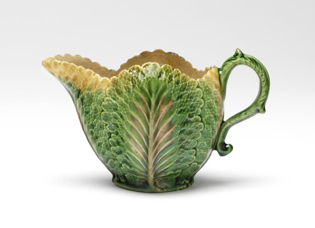 An image of Sauce boat/sauceboat. Unidentified Staffordshire Pottery. Moulded with overlapping leaves and decorated with coloured glazes. The boat stands on an oval base with slightly pointed ends. The sides are moulded with cabbage leaves with serrated edges at the top of the sides and lip. The ear-shaped handle is moulded with a short leaf at the top which forms the thumb piece, and two longer leaves down its back, the lower one having a kick at the bottom, and is glazed green overall. Dark cream earthenware, moulded in two halves, with an applied moulded handle, and decorated with manganese-brown oxide colour, and green, yellow and clear lead-glazes. Part of the base is unglazed, and there is an unglazed area on one side of the interior, possibly a finger mark. Height, whole, 6.6 cm, length, whole, 12.2 cm, circa 1755-1765. Rococo. Sir Ivor and Lady Batchelor Bequest through The Art Fund.