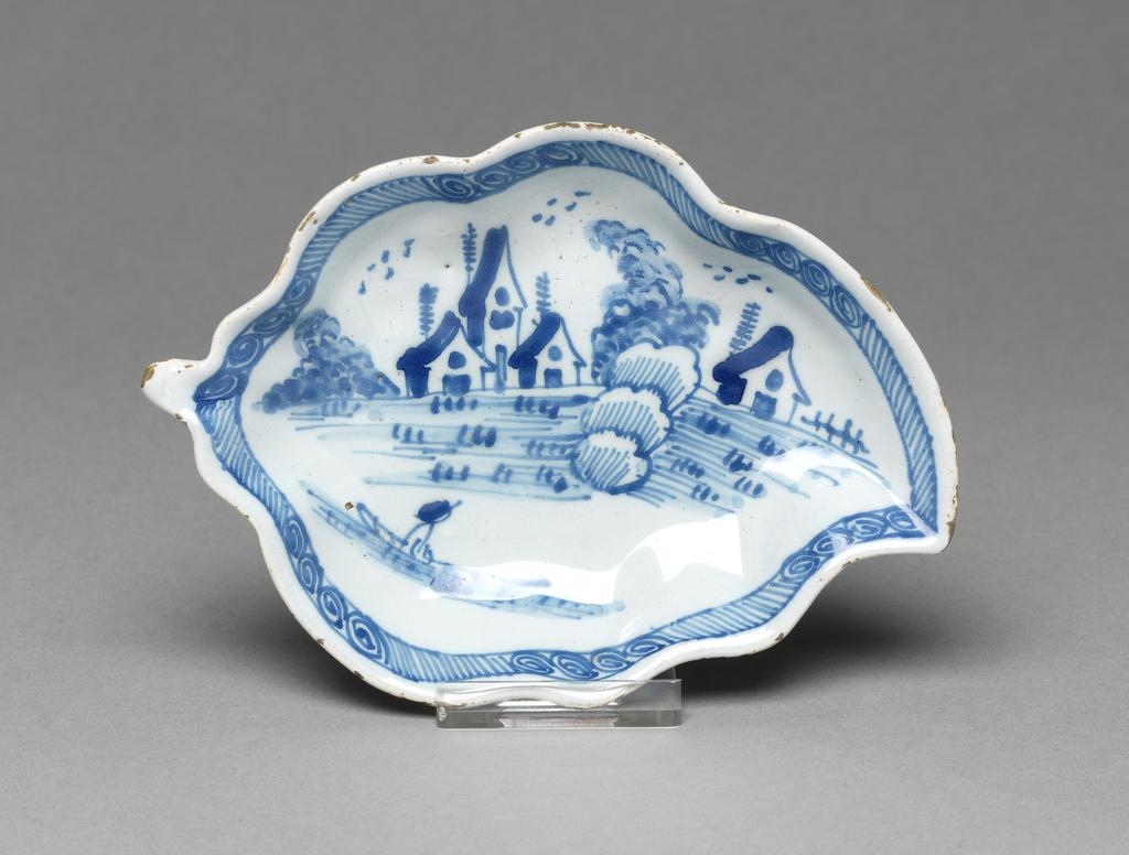 An image of English Delftware. Leaf dish. Pickle dish. Unidentified Liverpool pottery, England. Both dishes are moulded in the shape of a leaf with wavy edges and a short stalk, standing on a shaped footring. The interiors are painted with a lakeside scene in which there is a man in a boat in the foreground, an area with tufts of grass or reeds, three houses flanked by bushes and a single house with a fence to the right of it. Behind each house there is a slender tree resembling a telegraph pole, and overhead are three groups of schematic birds. The edge has a border of slanting lines broken by six groups of four spirals. Buff earthenware, press-moulded, tin-glazed pale bluish-white and painted in blue. Height, A, 2.2 cm, length, A, 13.2 cm, width, A, 9.2 cm; height, B, 2.2 cm, length, B, 13.1 cm, width, B, 9.3 cm, circa 1760. Rococo. Production Note: Small dishes of this shape were probably used for pickles presented on the table between the larger dishes in the course, or possible for sweetmeats for the dessert. Sir Ivor and Lady Batchelor Bequest through The Art Fund.