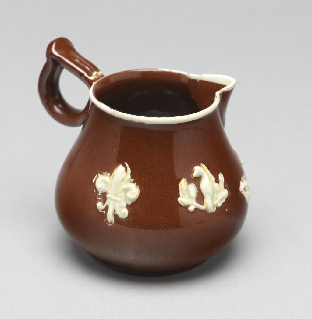 An image of Cream jug/milk jug. Unidentified Staffordshire Pottery, England. The bulbous body rises into a short cylindrical neck with a sparrow beak lip, and at right angles to it, a ring handle pinched together at the top. Below the lip is a sprig of bird perching on an upright branch with a large leaf on either side. Further to the left is a smaller bird perching on a branch with leaves, and to the left of that, a fleur-de-lys. A white slip band runs round the rim. Red earthenware, thrown, with applied spout and handle, decorated with mould-applied white sprigs (reliefs) and white slip under lead-glaze. Height, whole, 7 cm, width, whole, 8.4 cm, circa 1740-1750. Rococo. Sir Ivor and Lady Batchelor Bequest.