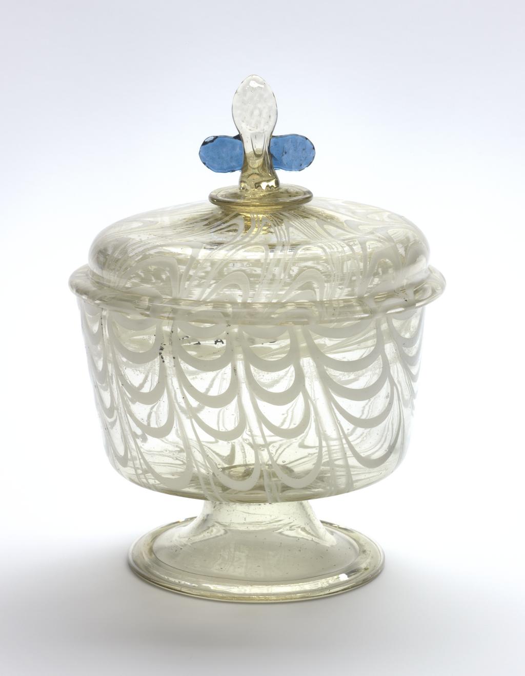 An image of Consecration bowl and cover. Unknown glasshouse, Spain. Clear glass, with opaque white combing. The cover has a pinched cross and two blue ears. Height 11 cm, diameter 8.7 cm, after 1550 to before 1650. Acquisition Credit: Sir Ivor and Lady Batchelor Bequest through The Art Fund.