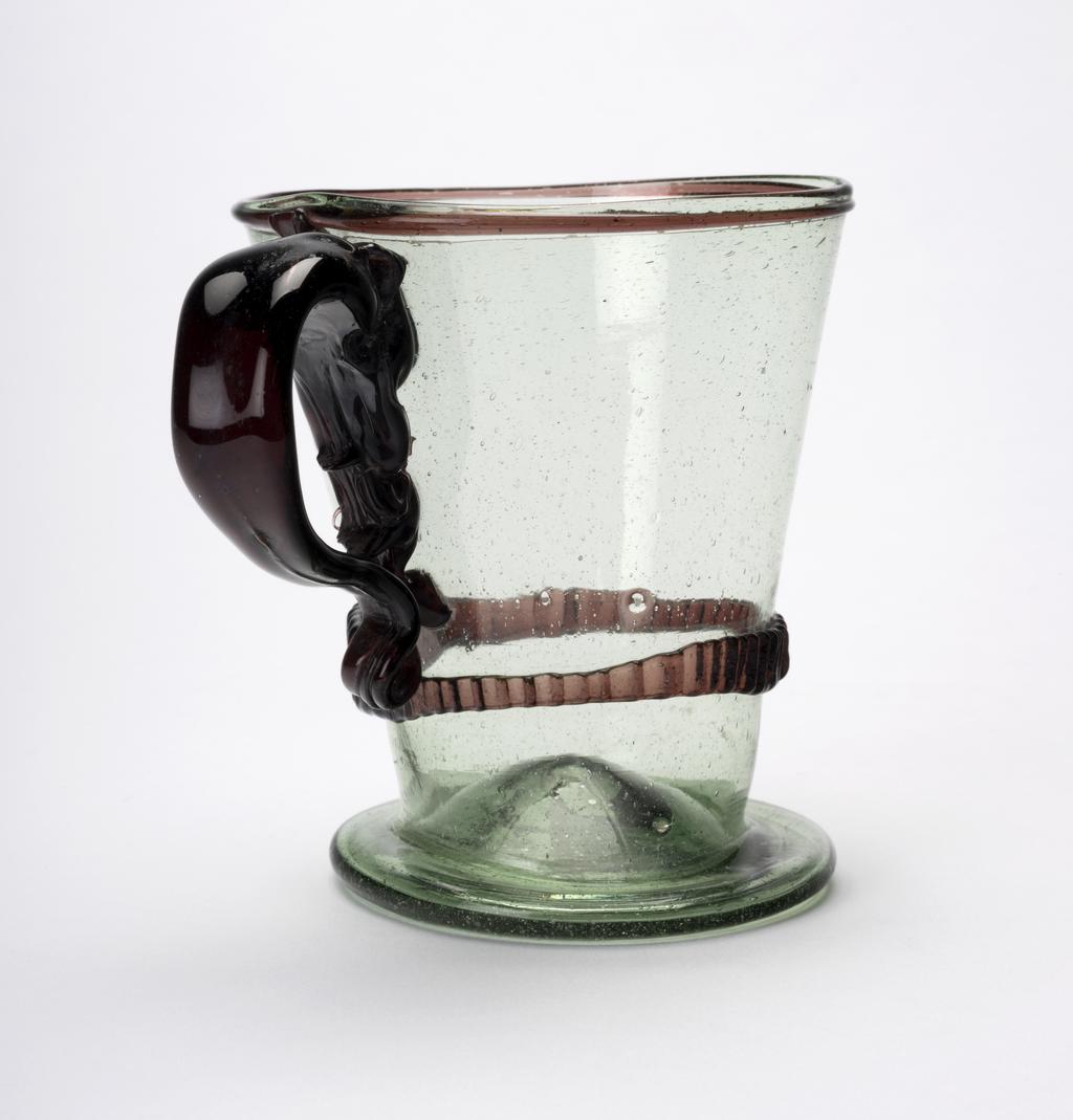 An image of Continental glass. Mug. Unknown glasshouse, Spain, Granada province. The mug has a projecting circular foot  with a pontil mark in the centre. The sides slope outwards to the rim which has an applied manganese-brown cord round its edge. A milled manganese-brown band of varying wide is applied a short distance above the foot. The handled is thicker and wider at the top than at its base which is curled under and extends upwards on the side of the mug to meet the top of the handle. Pale green glass, blown, with an applied manganese-brown cord, a milled brown band, and an applied brown handle. Height, whole, 10.4 cm, width, whole, 13.2 cm, diameter, rim, 9.3 cm, circa 1700-1800. Sir Ivor and Lady Batchelor Bequest.