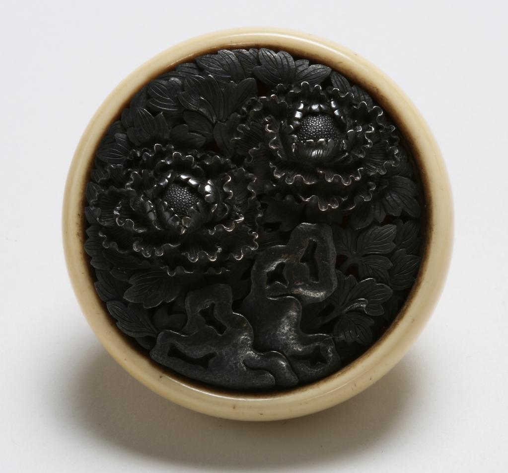 An image of Netsuke: kagamibuta: flowers. Unknown maker, Japan. Circular plain base with central himotoshi. Shibuichi disc cast in high relief and tooled in intricate detail with two blooming flowers set side by side; the whole disc is covered in leaves at different stages of relief. Holes punched through from the front are jagged on the underside. Ivory bowl with metal disc and eyelet held together by a brown shoelace; disc cast in high relief and tooled, height, whole, 1.9 cm, width, whole, 4.7 cm, circa 1800-1868. Edo Period (1615-1868).