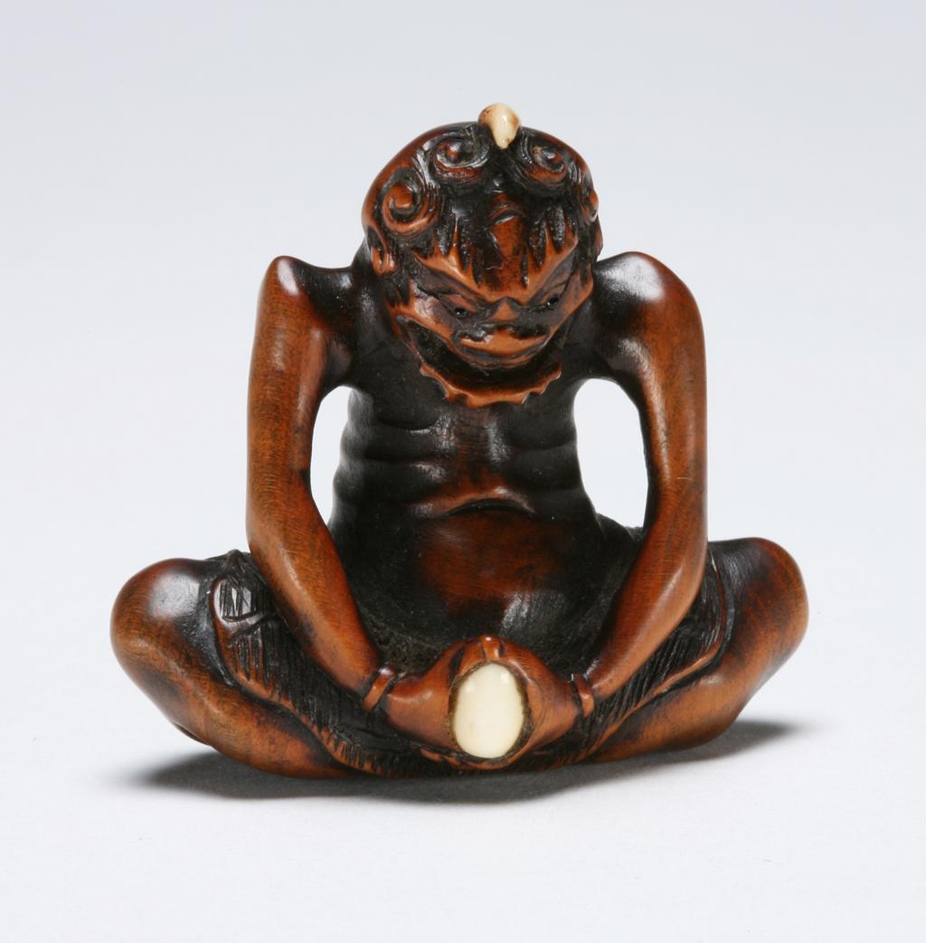 An image of Netsuke. Unknown maker, Japan. One horn Oni seated crossed legs. Ivory and horn inlaid on Tama (sacred jewel). Boxwood, carved, the eyes are inlaid with horn, 1800-1850.