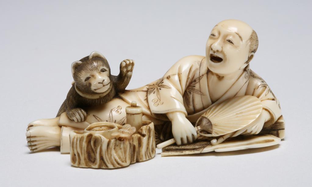 An image of Okimono. Unknown maker, Japan. A man reclining, resting his left arm on his books, while holding his pipe in his left hand and holding a fan in his right hand, he is pleased to see his pet cat on his legs, reaching out to the food container in the fore ground with its left paw. Ivory, 1870-1900.