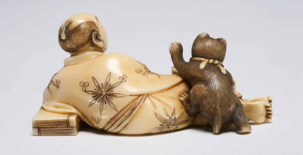 An image of Okimono. Unknown maker, Japan. A man reclining, resting his left arm on his books, while holding his pipe in his left hand and holding a fan in his right hand, he is pleased to see his pet cat on his legs, reaching out to the food container in the fore ground with its left paw. Ivory, 1870-1900.