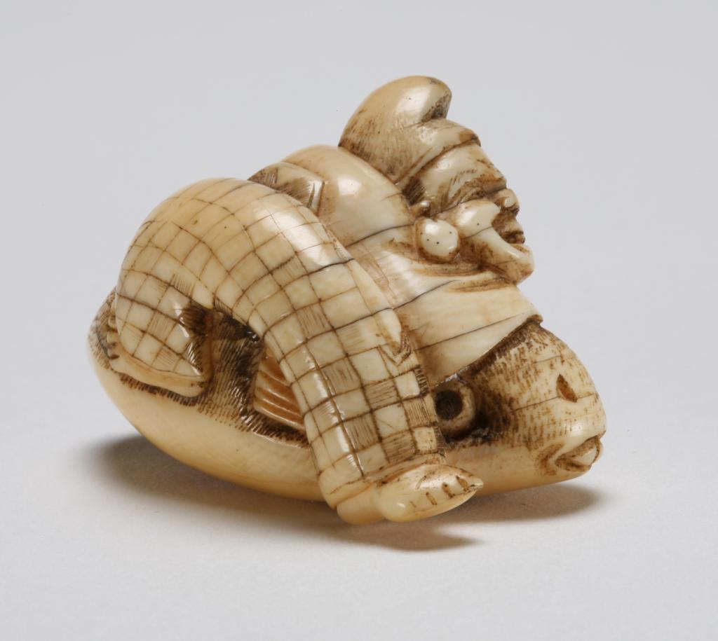 An image of Netsuke. Unknown maker, Japan. Ebisu is the protector of fisherman and merchants, he can be easily recognised by the court hat (Eboshi) and his fish. Here he tries to stop the large sea bream (Tai) moving away by leaning on top of the Tai. Ivory, the eyes of Tai-fish are inlaid with wood, 1800-1850.