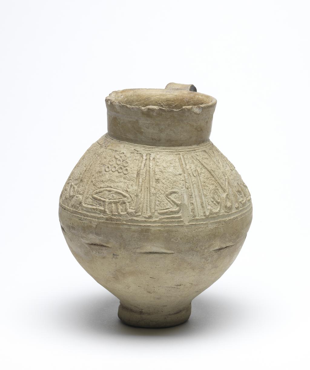An image of Islamic pottery. Jug. Unknown potter, probably, Iran or Syria. Shape: oval body on a disc base with a short, slightly flaring neck and a strap handle attached to the neck and shoulder. Exterior: a frieze of kufic script is moulded on the shoulder below which a series of impressed marks indicates the process of joining this decoration to the body of the jug. On the neck an impressed concentric band is visible. Interior: undecorated. Buff marl earthenware, probably wheel thrown with moulded decoration applied on the shoulder. Height, whole, 16.8 cm, width, whole, 14.7 cm, diameter, rim, 8.2 cm, diameter, base, 5.5 cm, weight, whole, 652 g, circa 1100-circa 1299. Ayyubid, Seljuk.