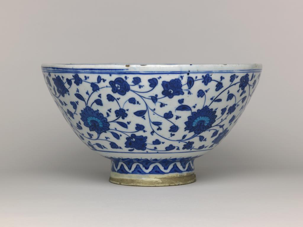 An image of Islamic pottery. Bowl. Unknown production, Turkey, Anatolia, Iznik. Shape: hemispherical bowl with a plain rim, sits on a tall, cylindrical foot ring. Interior: on the body a frieze of almond shaped medallions, containing rosettes and spiralling tadpole leaves reserved in turquoise, and flower pots containing symmetrical flower arrangements. On the base a central rosette with radiating tadpole leaves painted in a spiral and reserved in blue. Exterior: on the body, painted in blue, a frieze of large flowers, spiralling stems and leaves is painted above a band of angular, slanted leaves. On the foot stylised meandering scrollwork is reserved in blue. The base is glazed except for the rim of the foot ring. Fritware, probably wheel thrown, painted in blue and turquoise under a colourless glaze, height, whole, 15 cm, diameter, rim, 27.8 cm, diameter, base, 11 cm, circa 1530-circa 1535. Ottoman.