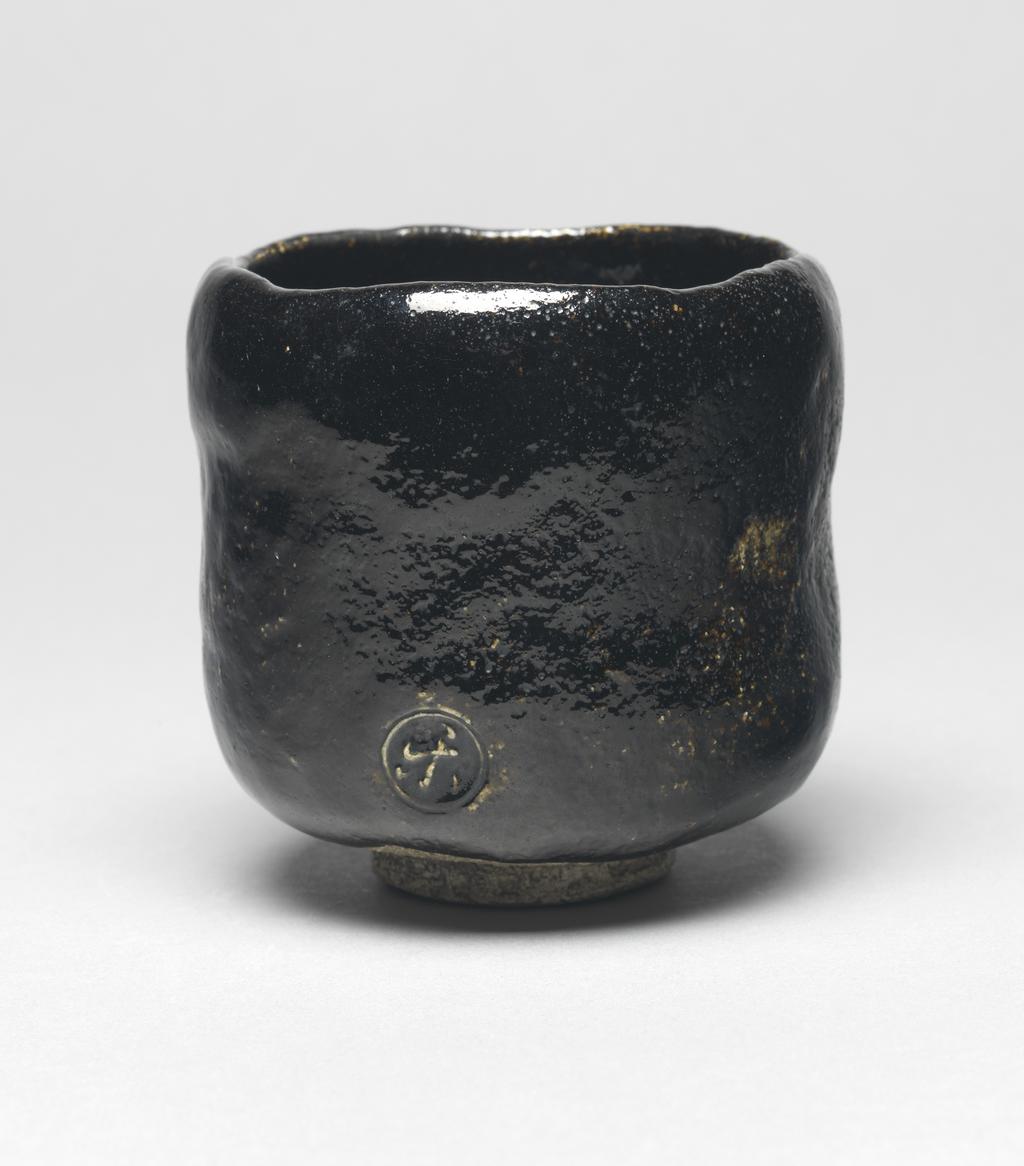 An image of Japanese pottery. Tea bowl. Ryonyu (Raku IX). Production Place: Japan, Kyoto. Hand-built footed tea bowl of very coarse grey-white earthenware with a thick black Raku glaze. Raku spiral in foot ring. Hollow at the centre of well. Vertical slicing on one side. Earthenware, glazed, height 9.40 cm, diameter, bowl, 10.00 cm, diameter, foot, 4.40 cm, circa 1775-circa 1825. Edo Period (1615-1868).