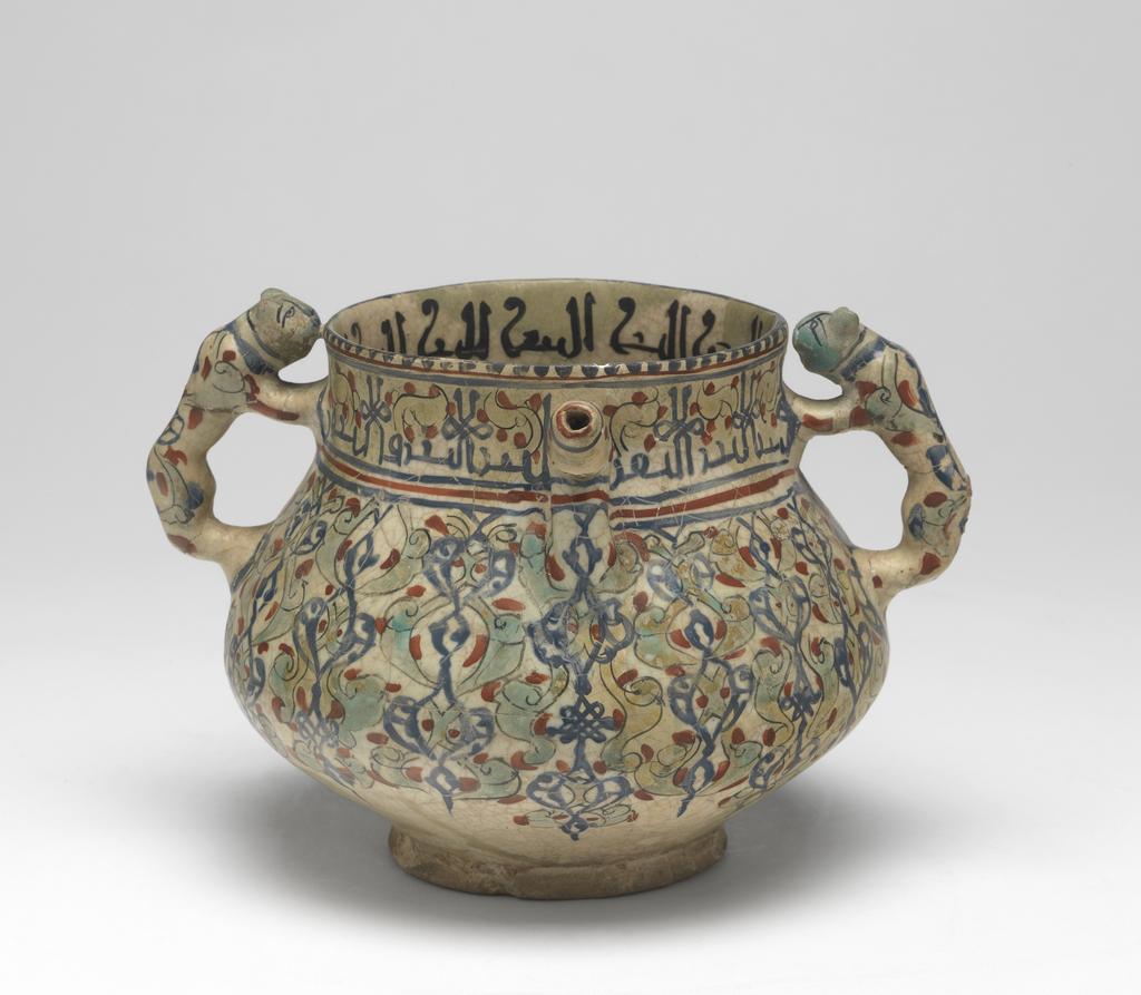 An image of Islamic pottery, Minai. Posset-pot. Unknown potter, Iran, central Asia, probably Kashan. Shape: bulbous body with a cylindrical neck and plain rim, sits on a low foot ring. Opposing cheetah handles and angular spouts are attached to the upper body. Exterior: on the rim a dentilated pattern is painted in blue. On the neck an epigraphic band is painted in blue and is interspersed with turquoise leaves and red dots. On the body arabesques of turquoise half-palmettes, sketchy blue birds and red dots covers the surface. On the cheetah handles and the spouts continuation of the patterns from the body are present, except on the head of the animals, which are painted turquoise with black lines to indicate features. Glaze covers the surface to the foot ring where it terminates unevenly. Interior: on the neck an epigraphic band is painted in black. The surface is coated in a glaze but otherwise is undecorated. Fritware, wheel thrown and mould-made, coated in white glaze painted with red, blue, turquoise and black. Height, whole, 14.6 cm, width, whole, 20.7 cm, diameter, rim, 11.4 cm, diameter, base, 8.7 cm, weight, whole, 694 g, circa 1170- circa 1220. Seljuk.