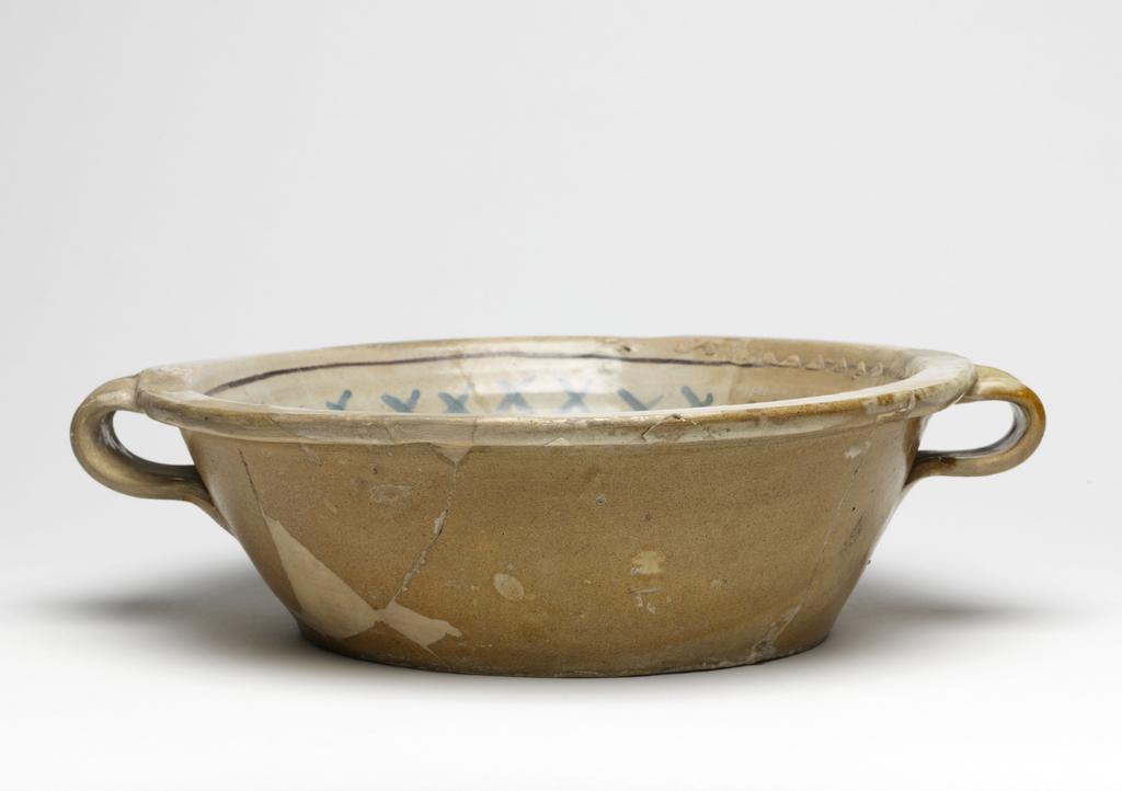 An image of Maiolica arcaica blu. Two-handled bowl. Unidentified pottery, probably Umbria, Orvieto or Tuscany. Shape 39. Circular with flat base, sloping sides, narrow flat rim and two wide strap handles with a thumb print at the top. Inside, within two manganese concentric circles, there is a double-armed cross with cross-hatching and three scalloped lines between the arms; on the sides, blue criss-crosses and a manganese band. Pale buff earthenware. The exterior and base are lead-glazed pale brown; the interior is tin-glazed pale grey. Painted in manganese and blue. Height, whole, 9.7 cm, diameter, base, 18.8 cm, width, whole, 34.0 cm, circa 1350-1425. Late Medieval.