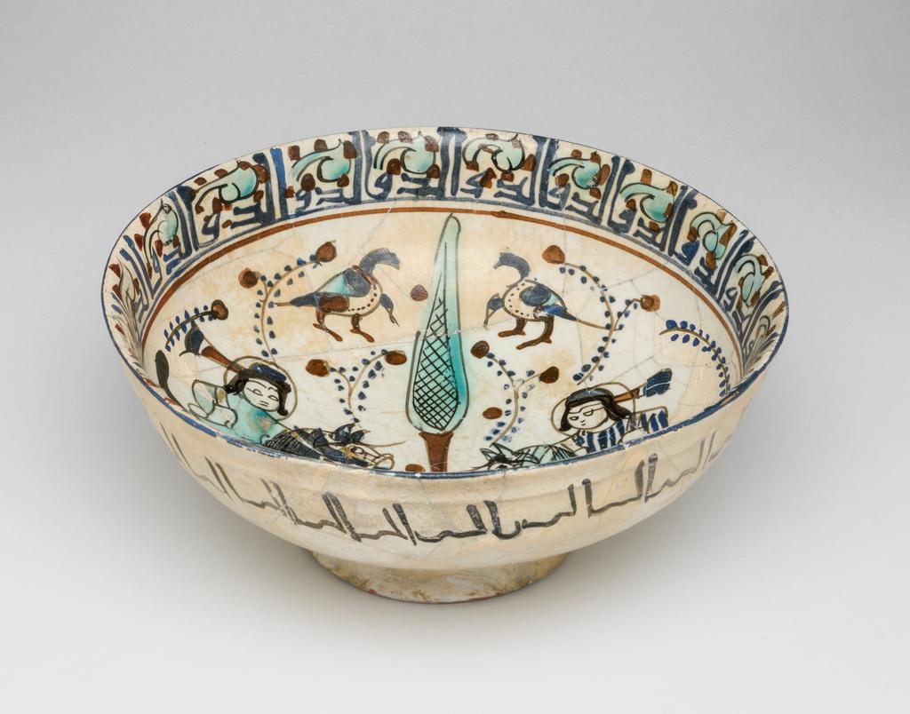 An image of Bowl. Islamic pottery. Minai. Unknown, potter, central Asia, probably Kashan. Shape: carinated bowl with collared neck, sits on a slightly flaring foot ring base. Interior: on the neck a kufic inscription is painted in blue and surrounded by turquoise leaves and red dots. On the body two mounted horsemen, wearing headdresses and tunics with gilding, are confronted either side of a cypress tree surrounded by leafy fronds dotes with blue and red. Above and below the horsemen pairs polychrome birds are painted. Exterior: on the upper body a cursive inscription is painted in black. Glaze covers the surface unevenly, terminating unevenly on the foot ring. Fritware, wheel thrown, coated in white glaze painted with dark red, blue, turquoise, black and gilded with gold leaf,. Height, whole, 7.4 cm, width, whole, 16.8 cm, diameter, rim, 16.8 cm, diameter, base, 7.2 cm, weight, whole, 242 g, circa 1170- circa 1220. Seljuk Period.