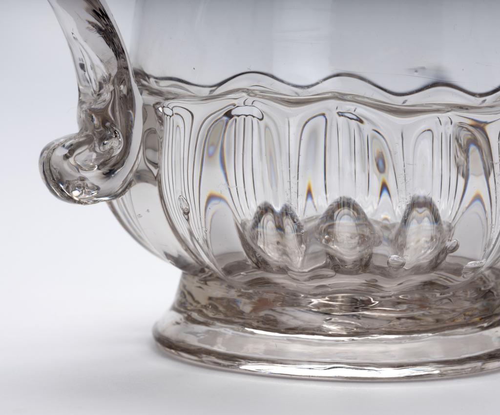 An image of Two-handled cup. Loving cup. Unidentified glasshouse, England. The thistle-shaped bowl has moulded vertical gadrooning round the lower part and stands on a low foot. It has an applied trailed thread about half way up the side. On each side it has an applied strap handle with an upward kick at the lower end. In the middle of the underside there is a pontil mark. Clear lead-glass, height, whole, 11.9 cm, width, whole, 16.5 cm, diameter, foot, 7.1 cm, 1710-1720. Baroque. Sir Ivor and Lady Batchelor Bequest through The Art Fund.