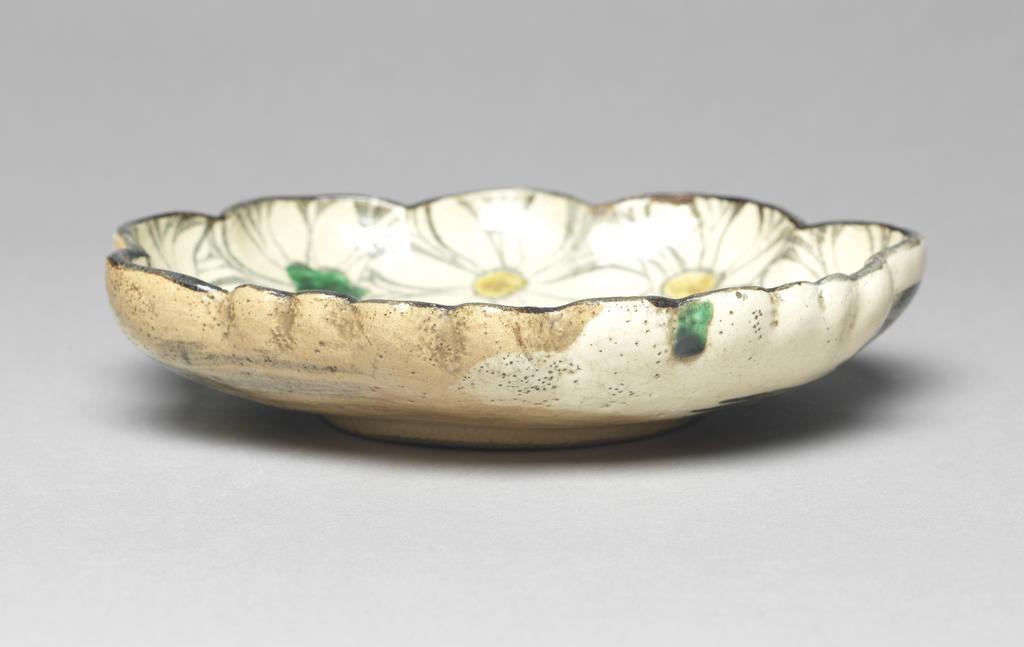 An image of Japanese pottery. Dish. Kenzan, Ogata (1663 - 1743) Japan, Kyoto. Light brown dish of foliate form, moulded with thrown foot. Fawn base glaze, then overglaze decoration with chrysanthemum flowers and foliage in white, yellow and green with black outlines. Some gold background. Stoneware, brush painted, height, whole, 3.30 cm, diameter, dish, 16.60, cm, largest, diameter, foot, 7.90 cm, circa 1600-1743. Edo Period (1615-1868).